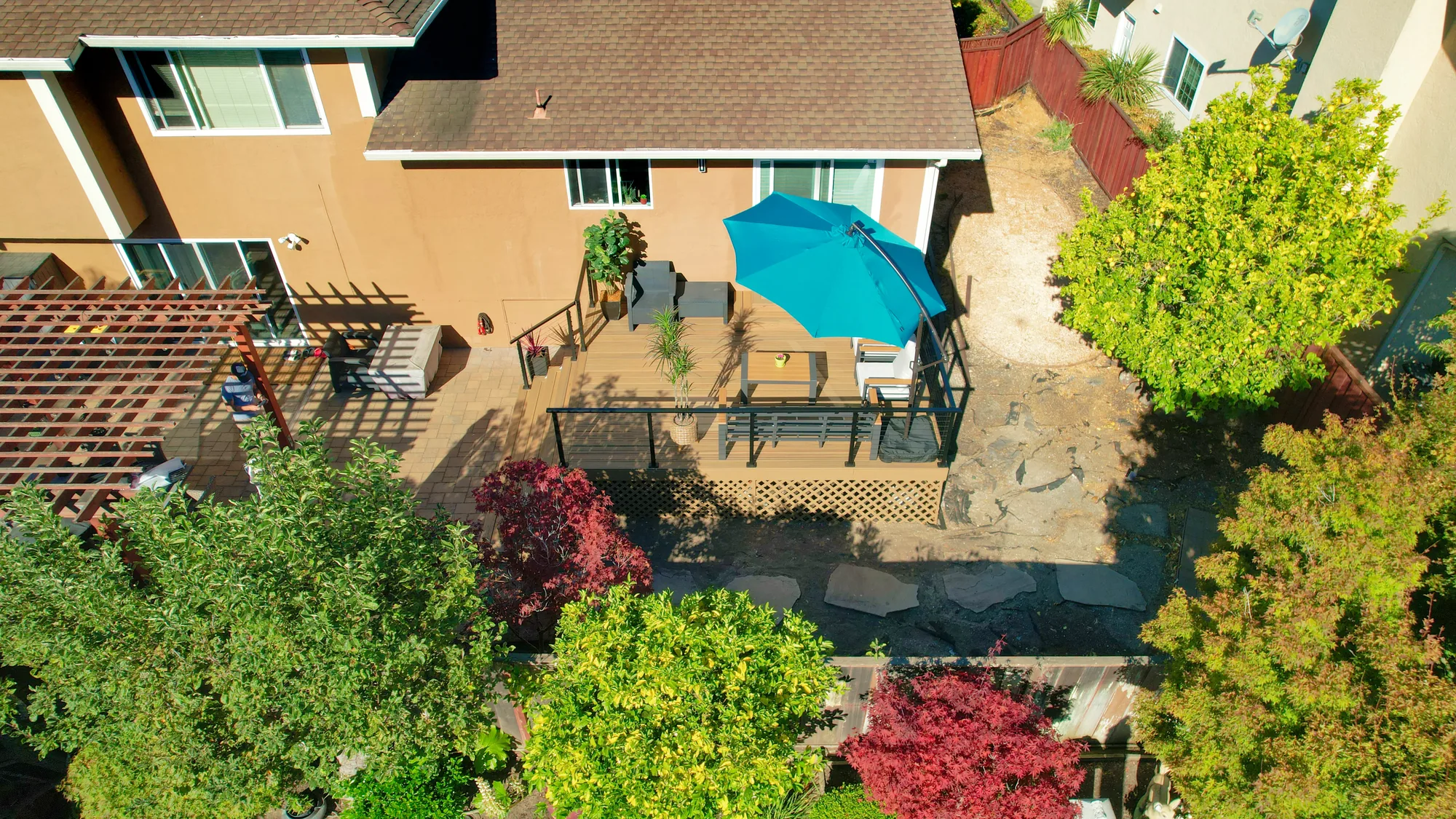 Aerial view of a backyard with a raised deck, pergola, paver patio and flagstone path among maple trees.