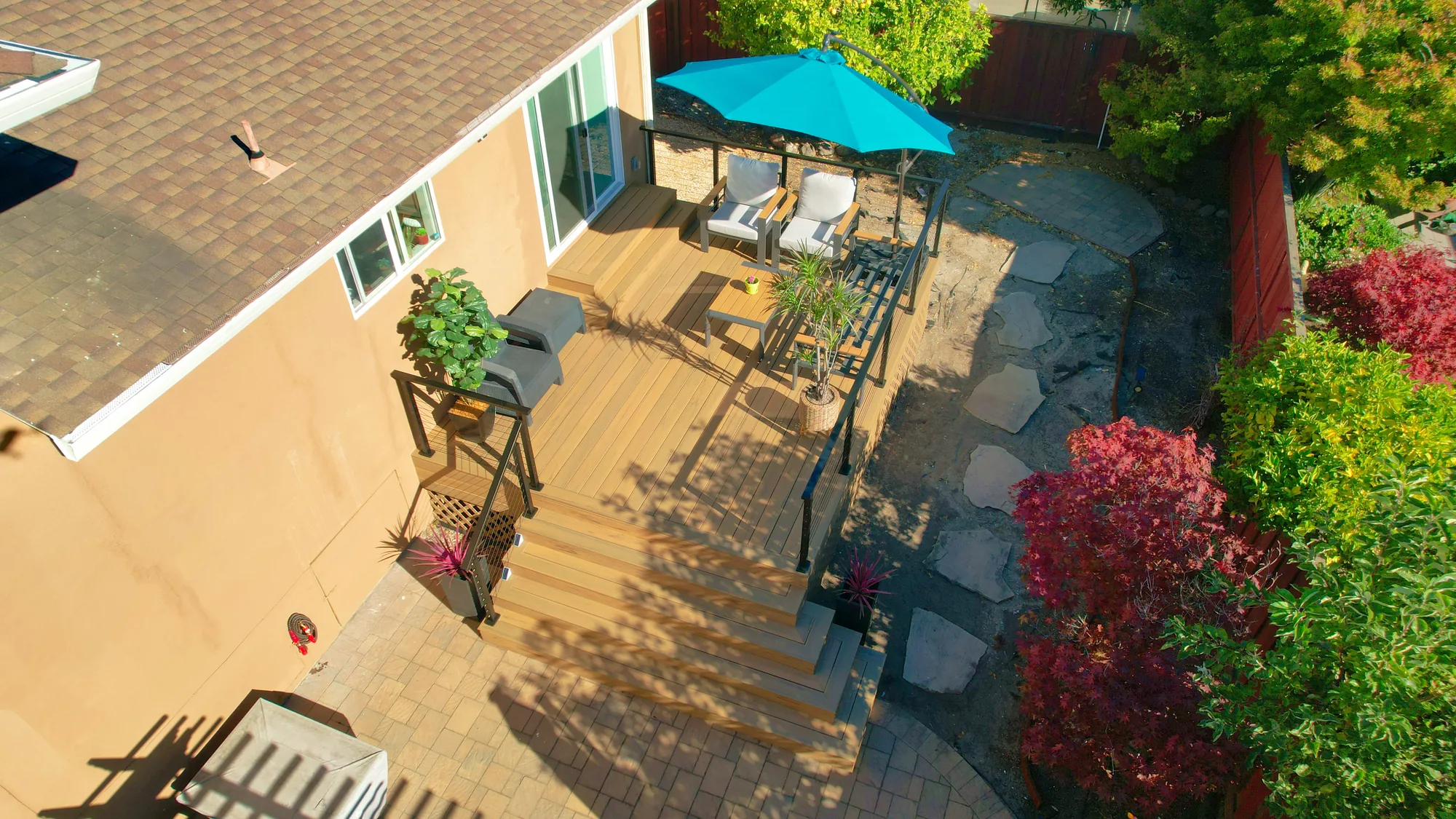 Angled aerial view of a tan deck with black cable railing and wide steps down to a paver patio.