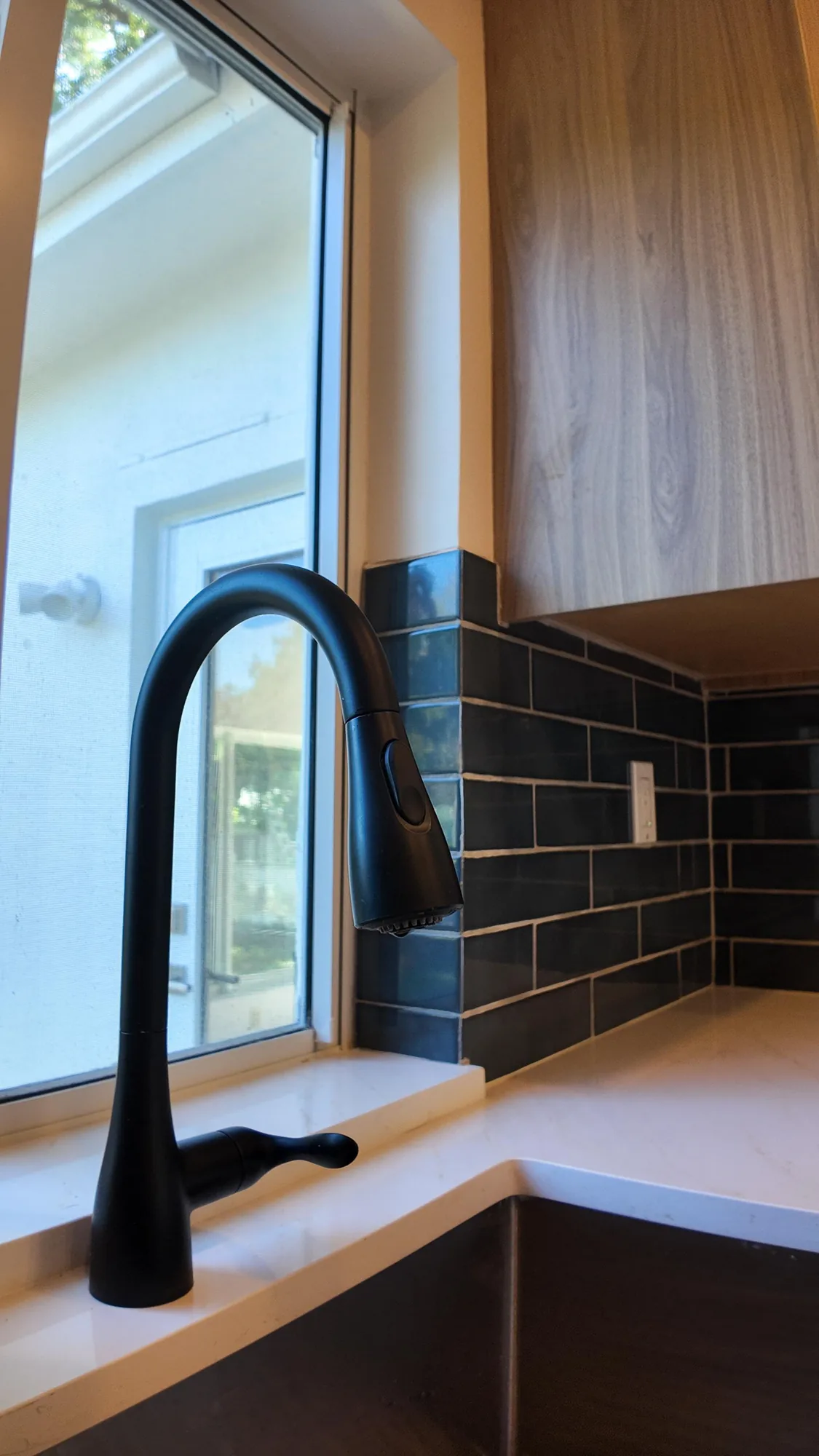 Matte black faucet at a kitchen window, with quartz sill and dark subway tile wrapping the corner.