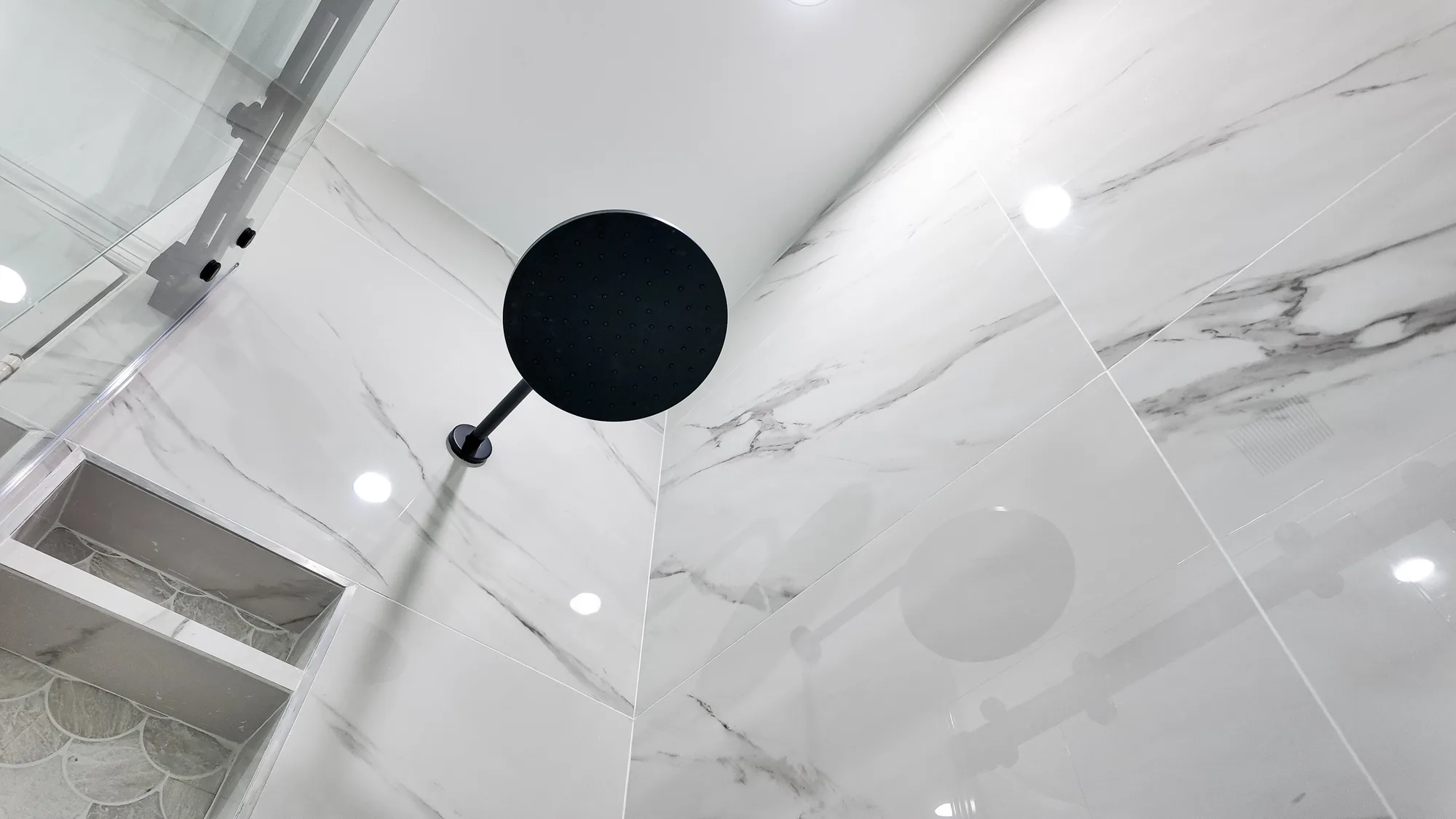 Upward view of a round matte black rain shower head and recessed ceiling lights over tiled walls.