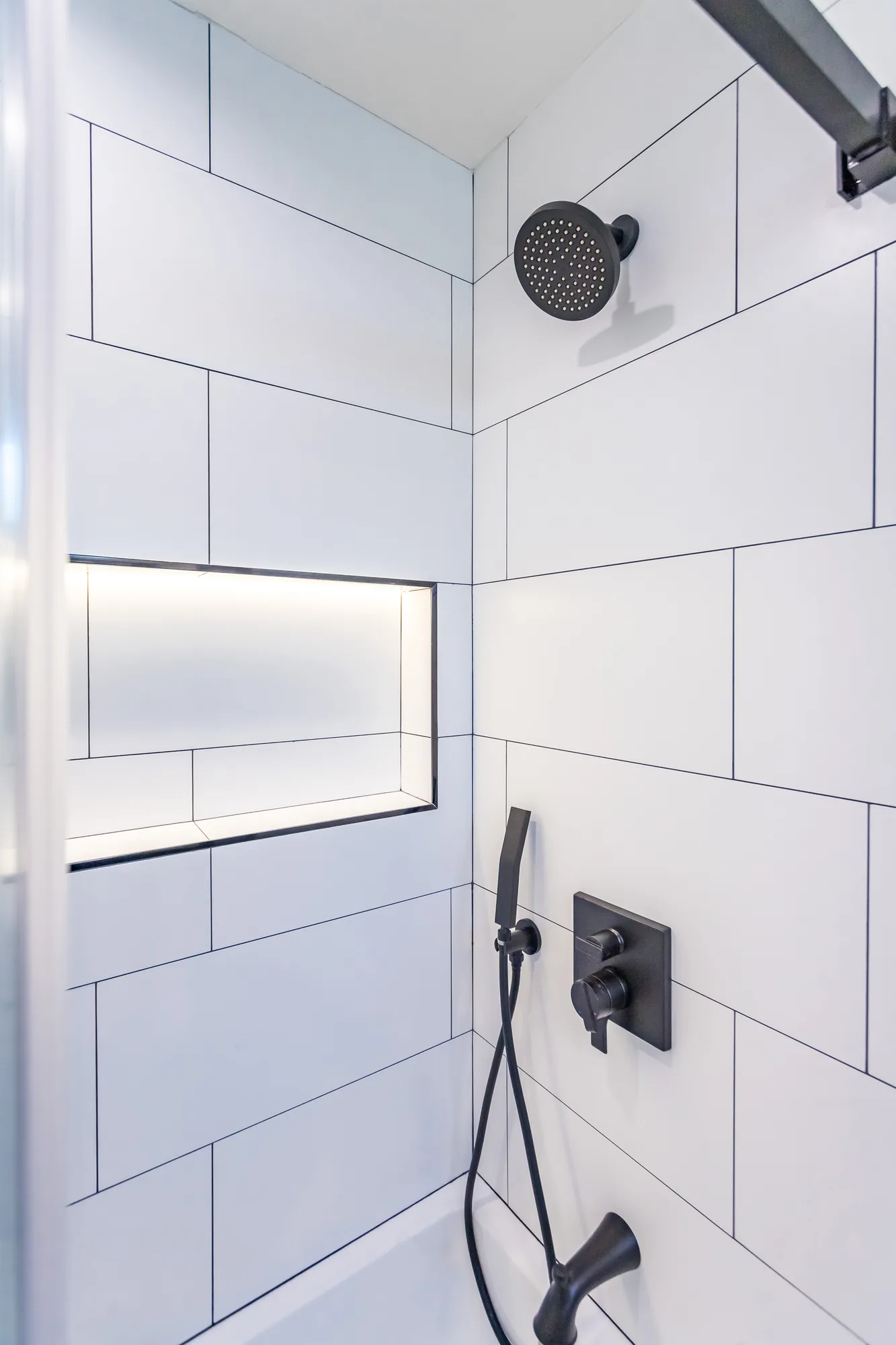 Shower interior showing a matte black showerhead, handheld sprayer and an illuminated tile niche.