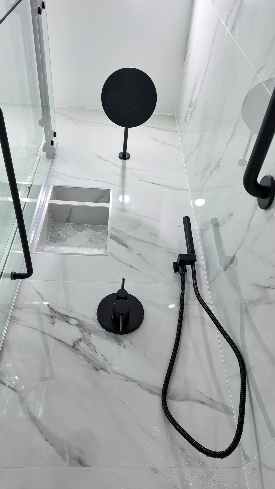 Matte black shower valve, handheld sprayer and rain head on marble-look tile inside a glass shower.