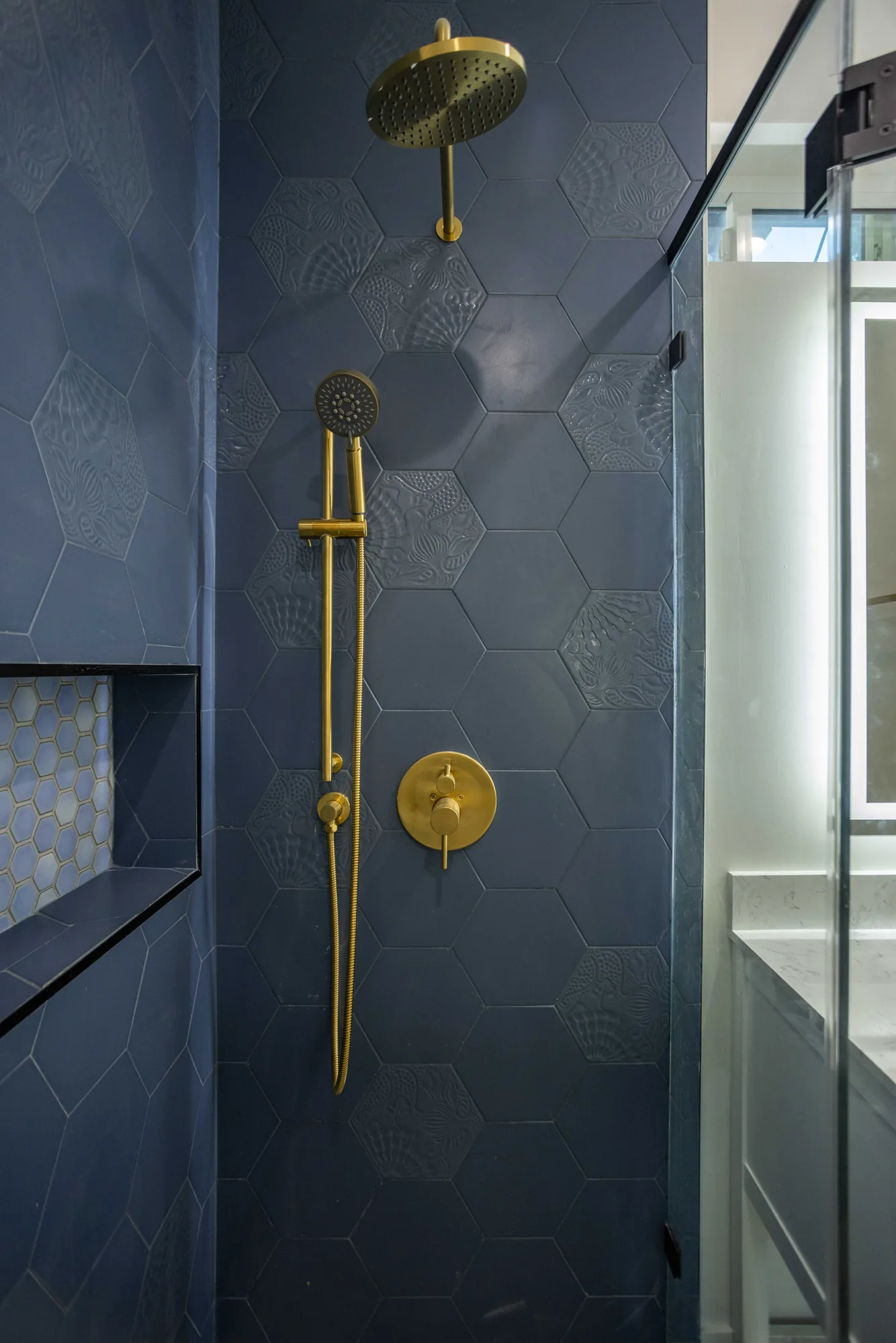 Brass rain shower head, handheld slide bar and round valve trim mounted on navy hexagon tile.