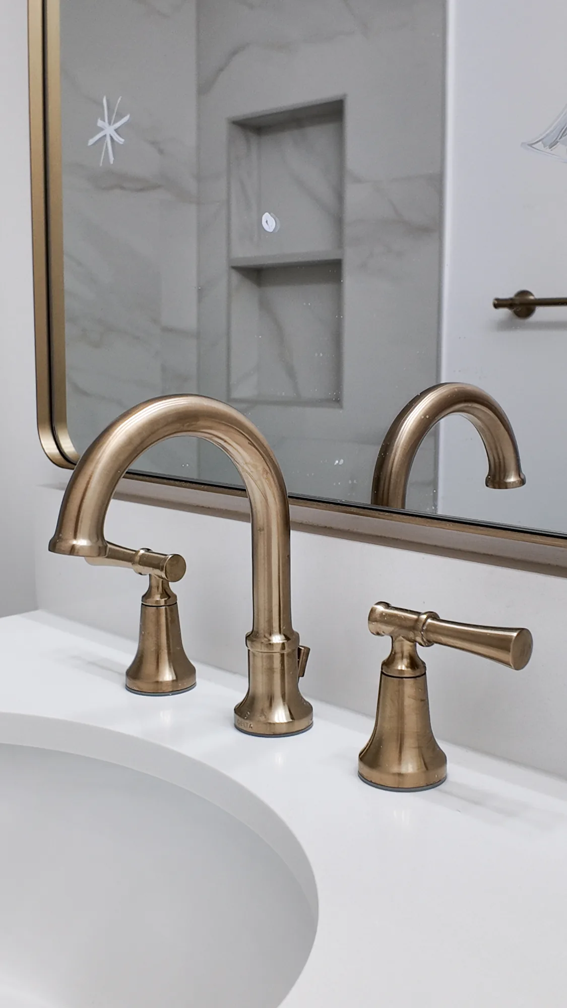 Brushed gold widespread faucet on a white vanity top, with a gold-framed mirror behind it.