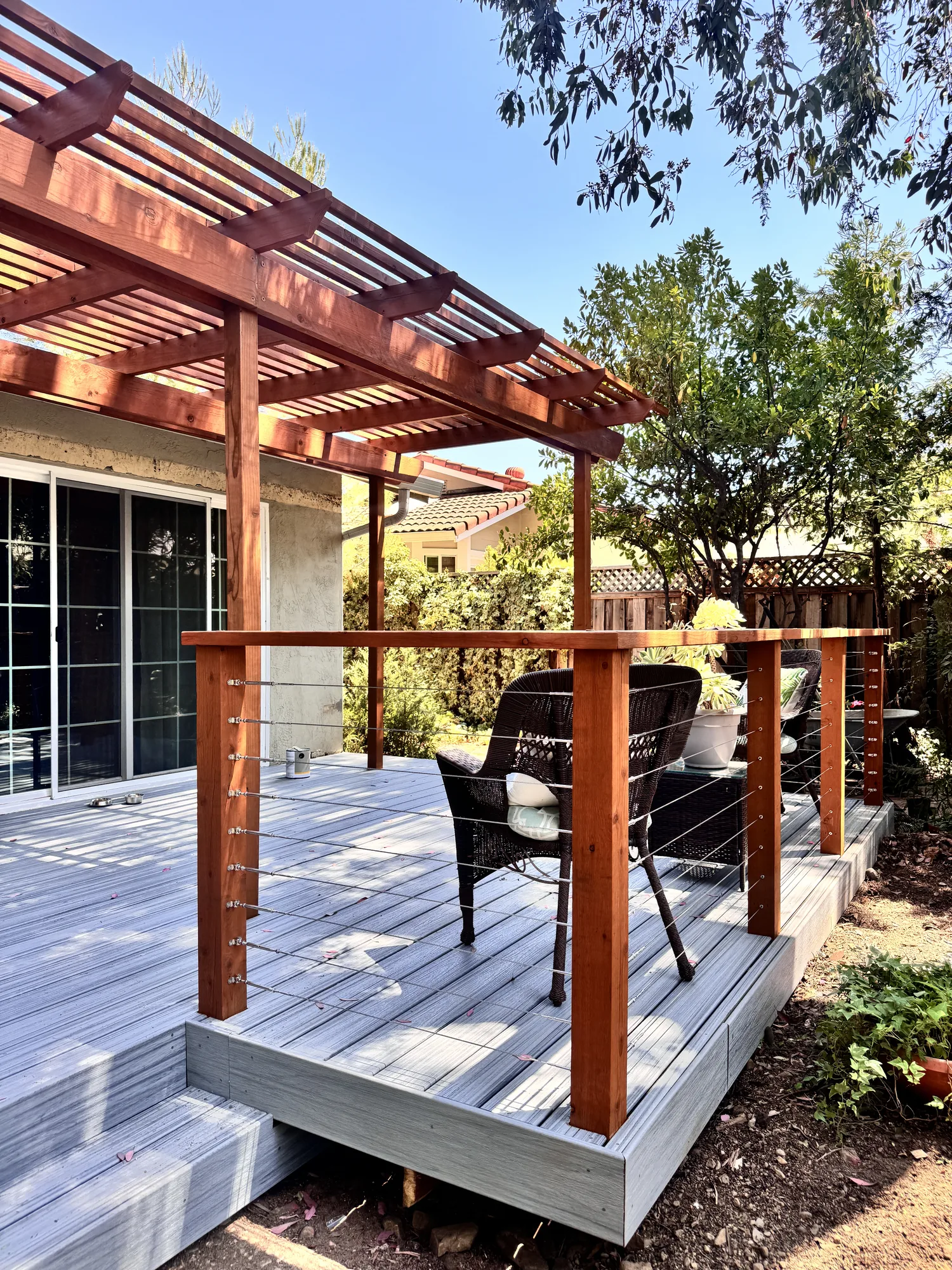 Deck seating area enclosed by a stainless cable railing with a pergola overhead and yard beyond.