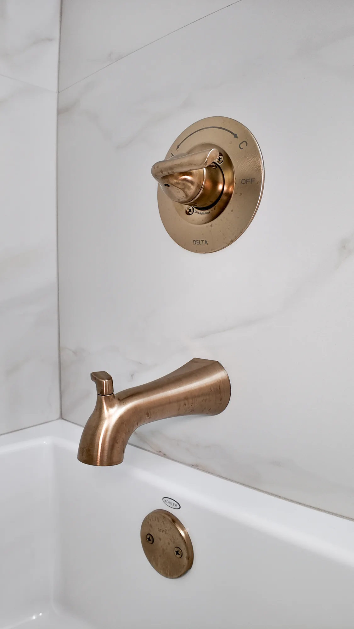 Champagne bronze tub spout and shower valve trim on marble-look tile above a white bathtub.