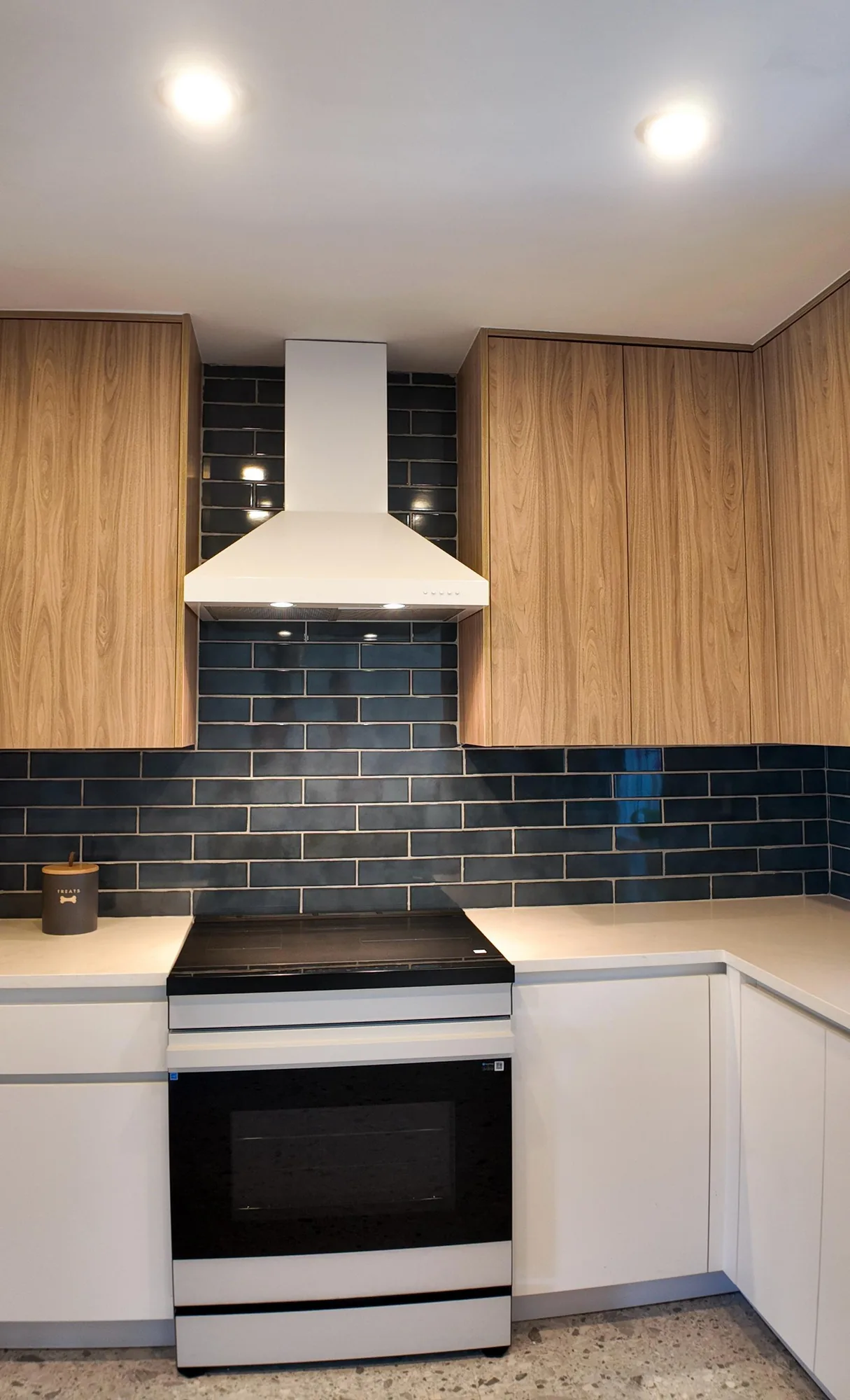White chimney range hood lit above a smooth-top range, with glossy dark tile filling the wall behind.