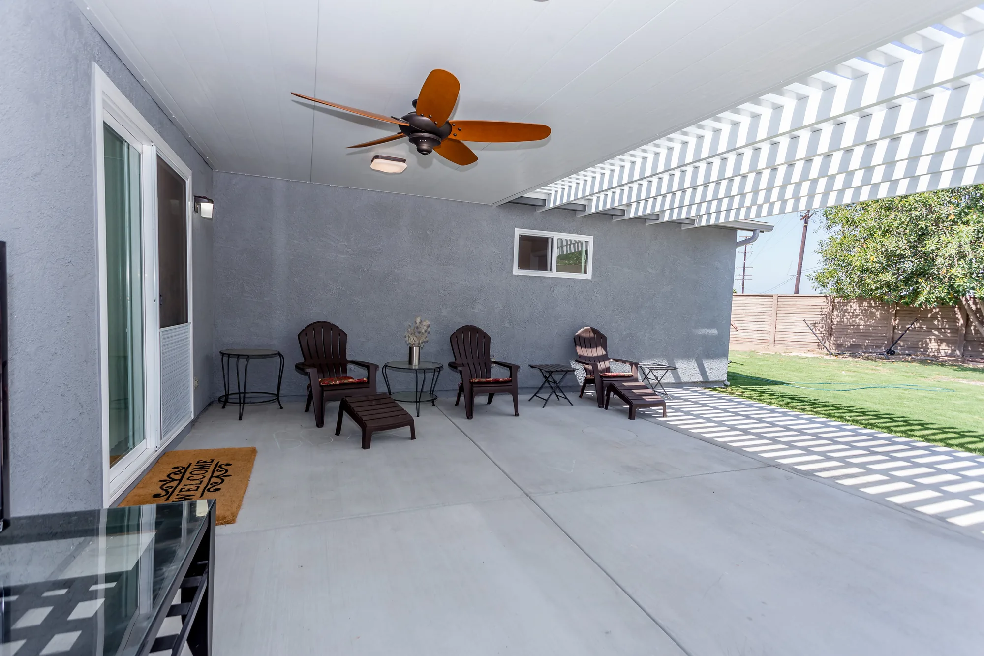 Covered patio with a wood-blade ceiling fan, gray stucco walls and a white pergola opening onto a lawn.