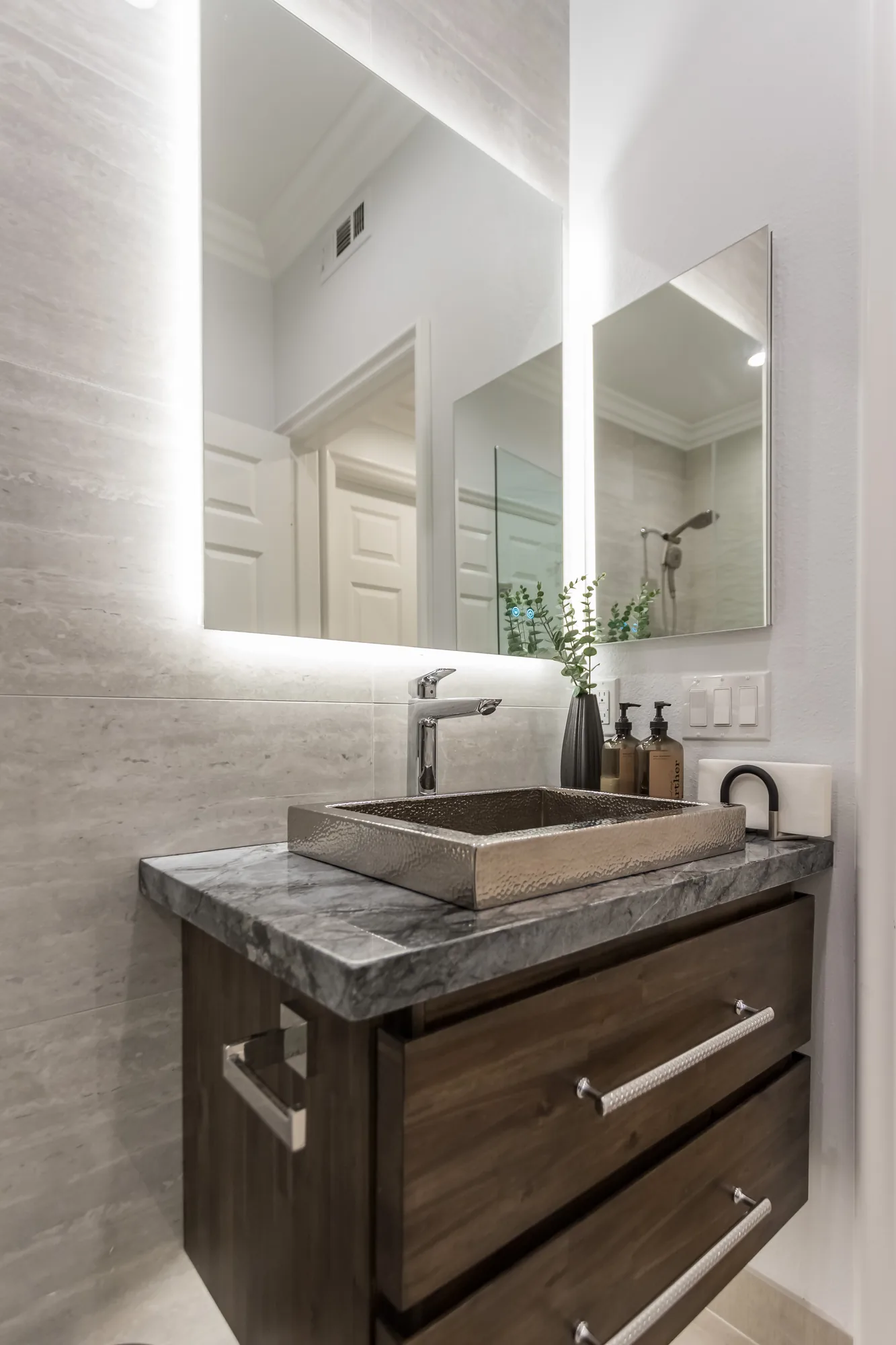 Dark wood vanity with a grey stone top, hammered metal sink and a backlit LED mirror on a tiled wall.