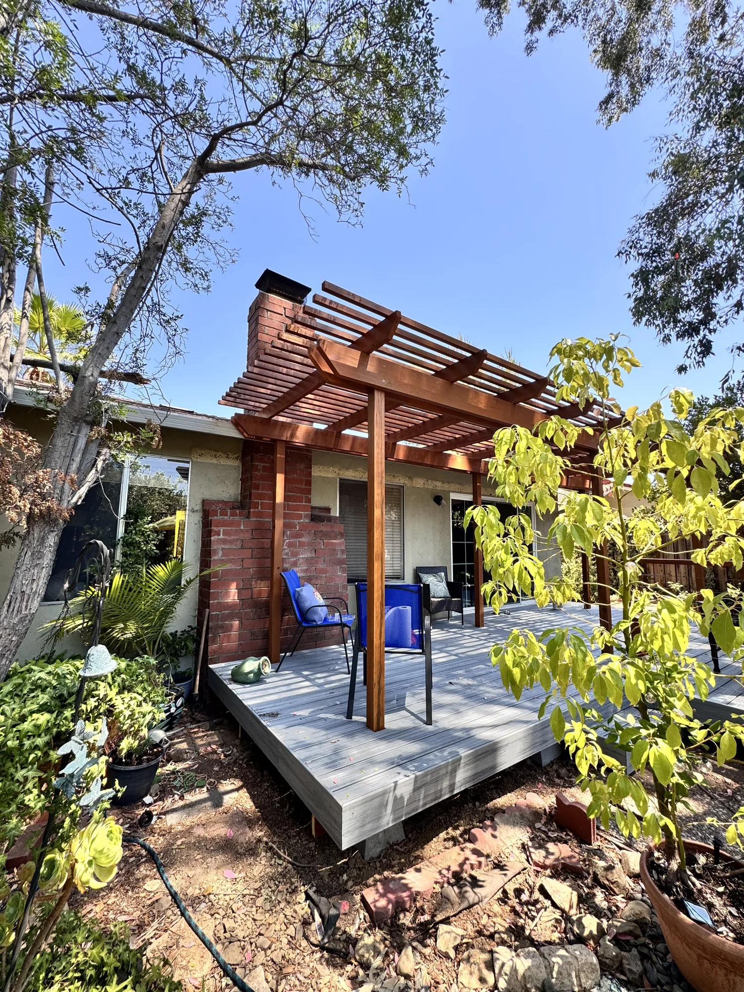 Corner of a raised gray deck with a stained wood pergola beside a brick chimney and garden plantings.