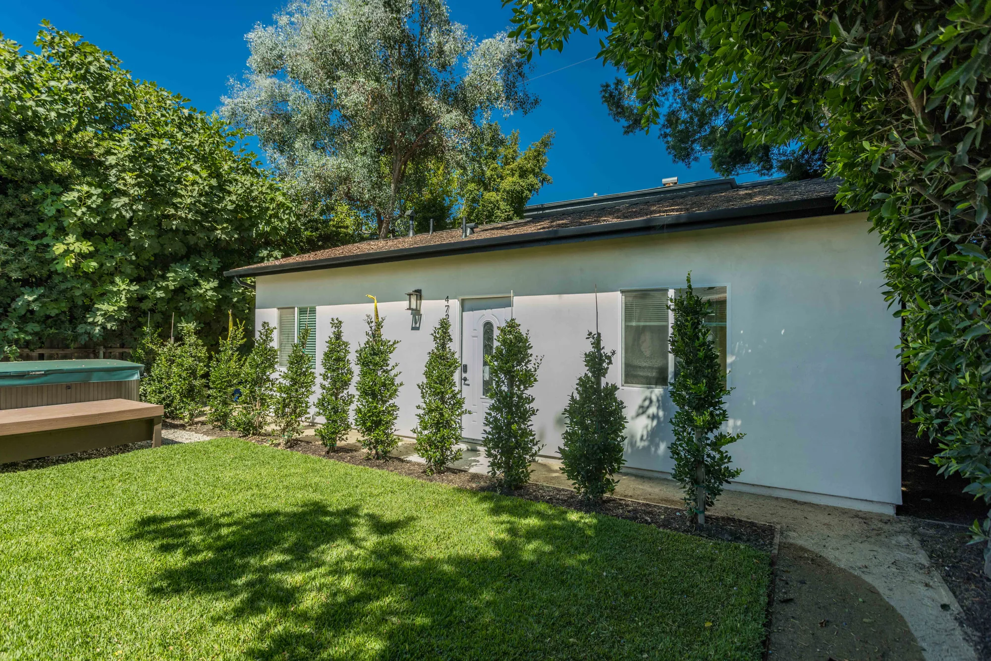 White stucco detached unit with a shingle roof, new hedge planting, lawn and a deck to one side.