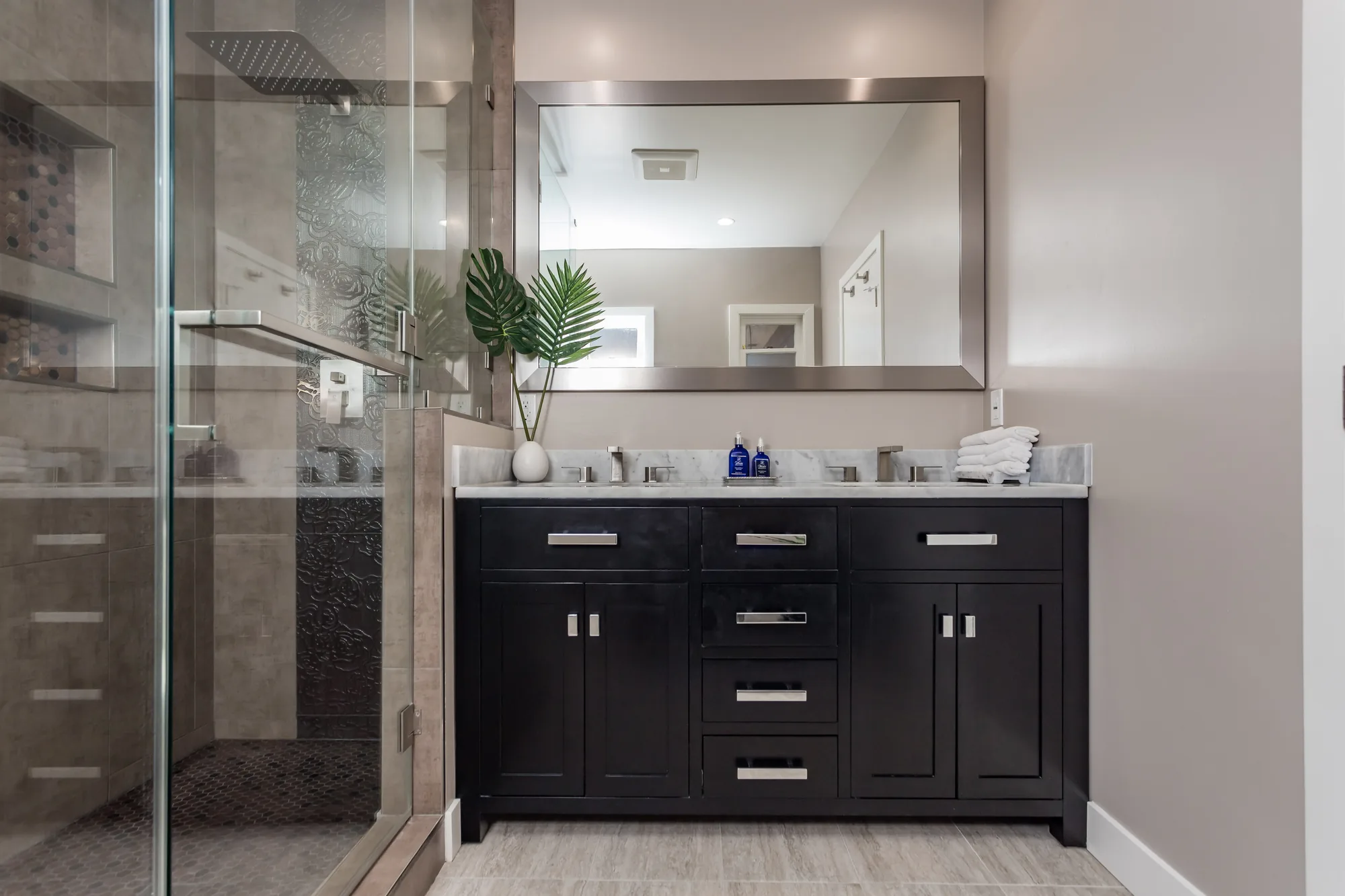 Dark double vanity with a stone-look counter and framed mirror beside a glass shower enclosure.