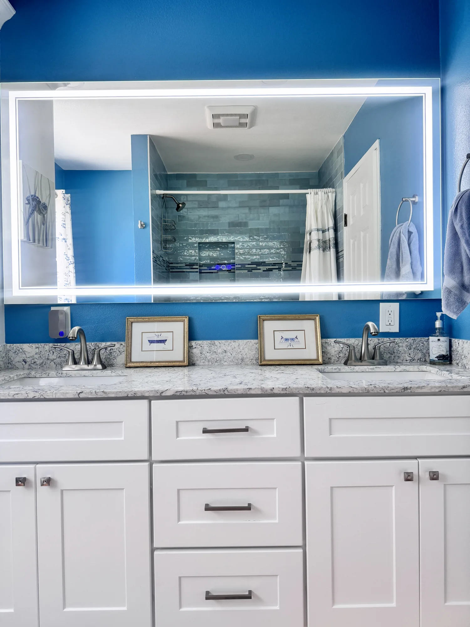 White shaker double vanity with veined stone-look counter and a large backlit mirror on a blue wall.