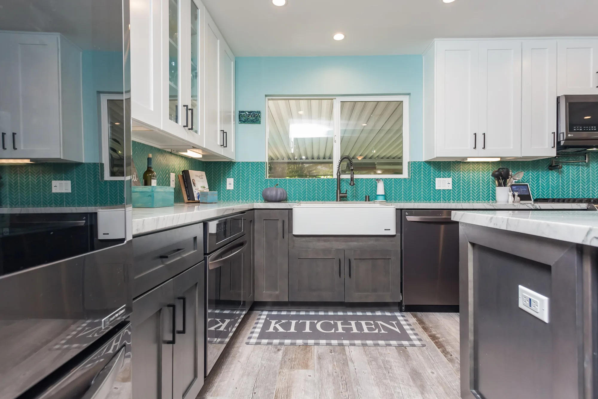 Farmhouse sink under a window with gray base cabinets, dishwasher, and teal tile backsplash.