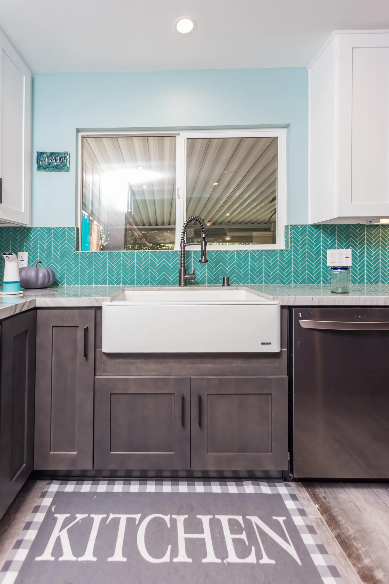 White farmhouse sink and window flanked by teal tile, gray cabinets, and a stainless dishwasher.