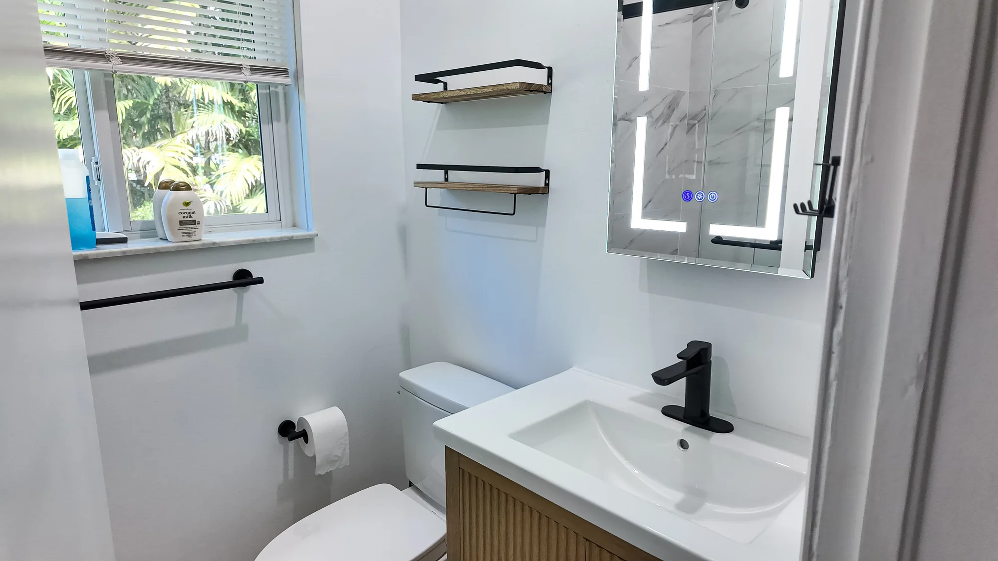 Wide view of a finished bathroom with a window, toilet, wood vanity and lighted mirrored cabinet.