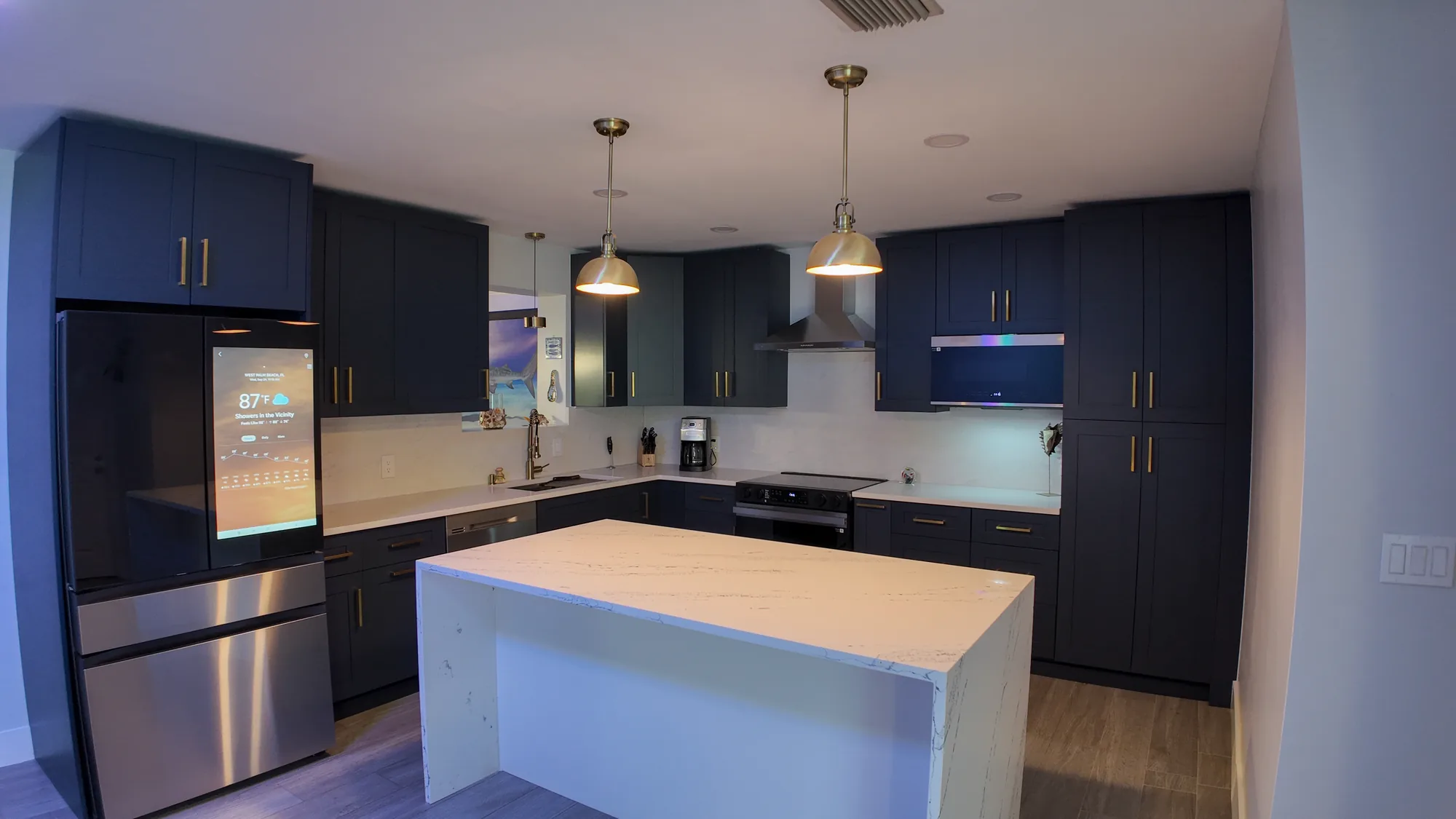 Wide view of a finished navy kitchen with a white quartz island, brass pendants and stainless appliances.