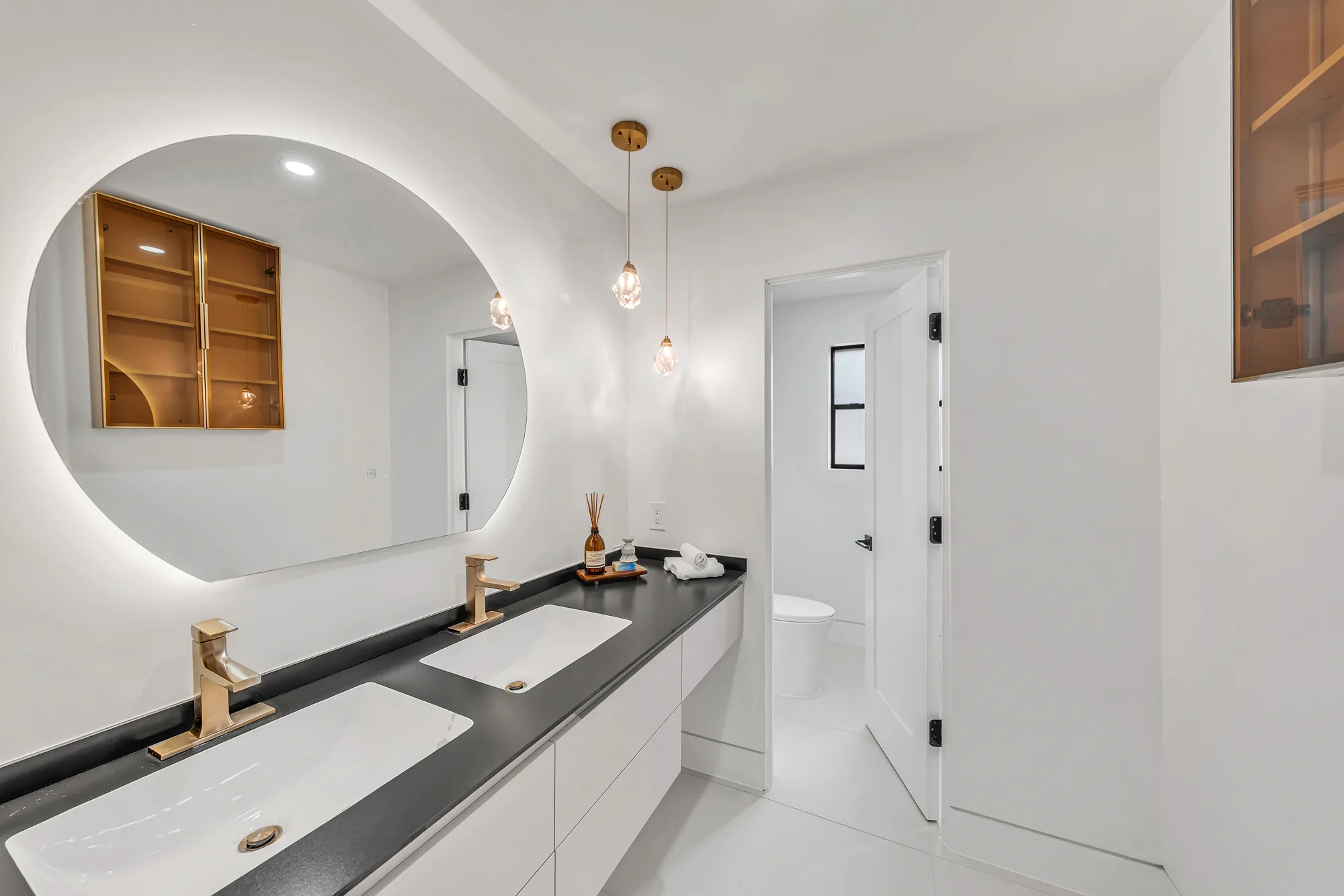 Floating white double vanity with dark stone counter, round backlit mirror and gold glass pendant lights.