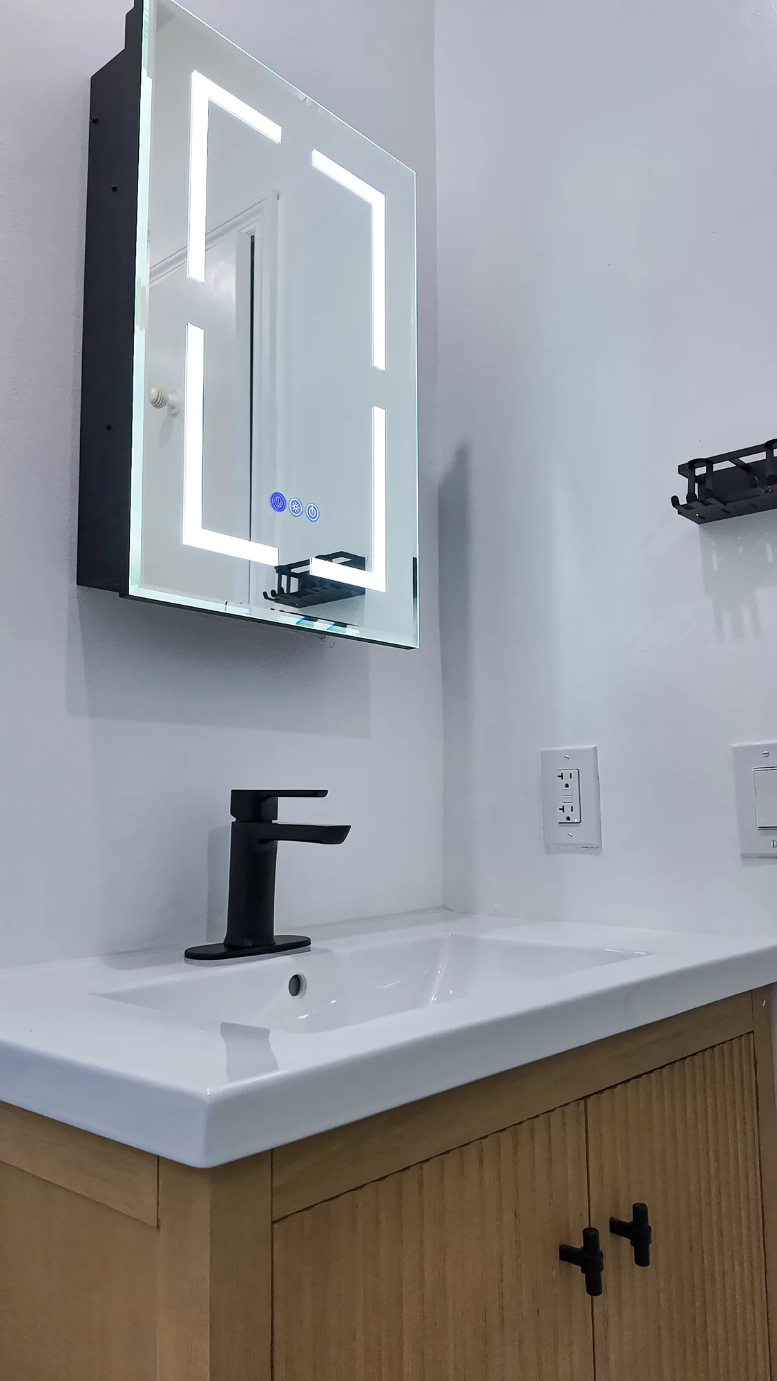 Fluted wood vanity with a white integrated sink, black faucet and a lighted mirrored medicine cabinet.