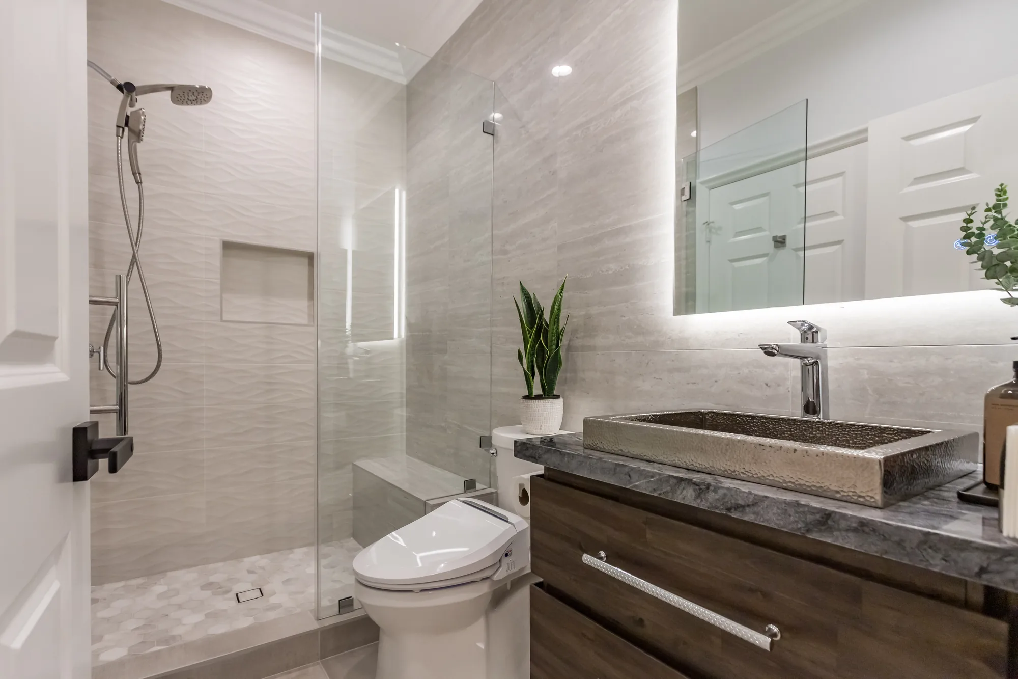 Wide view of a remodeled bathroom with a walk-in glass shower, dark wood vanity and stone-look tile walls.