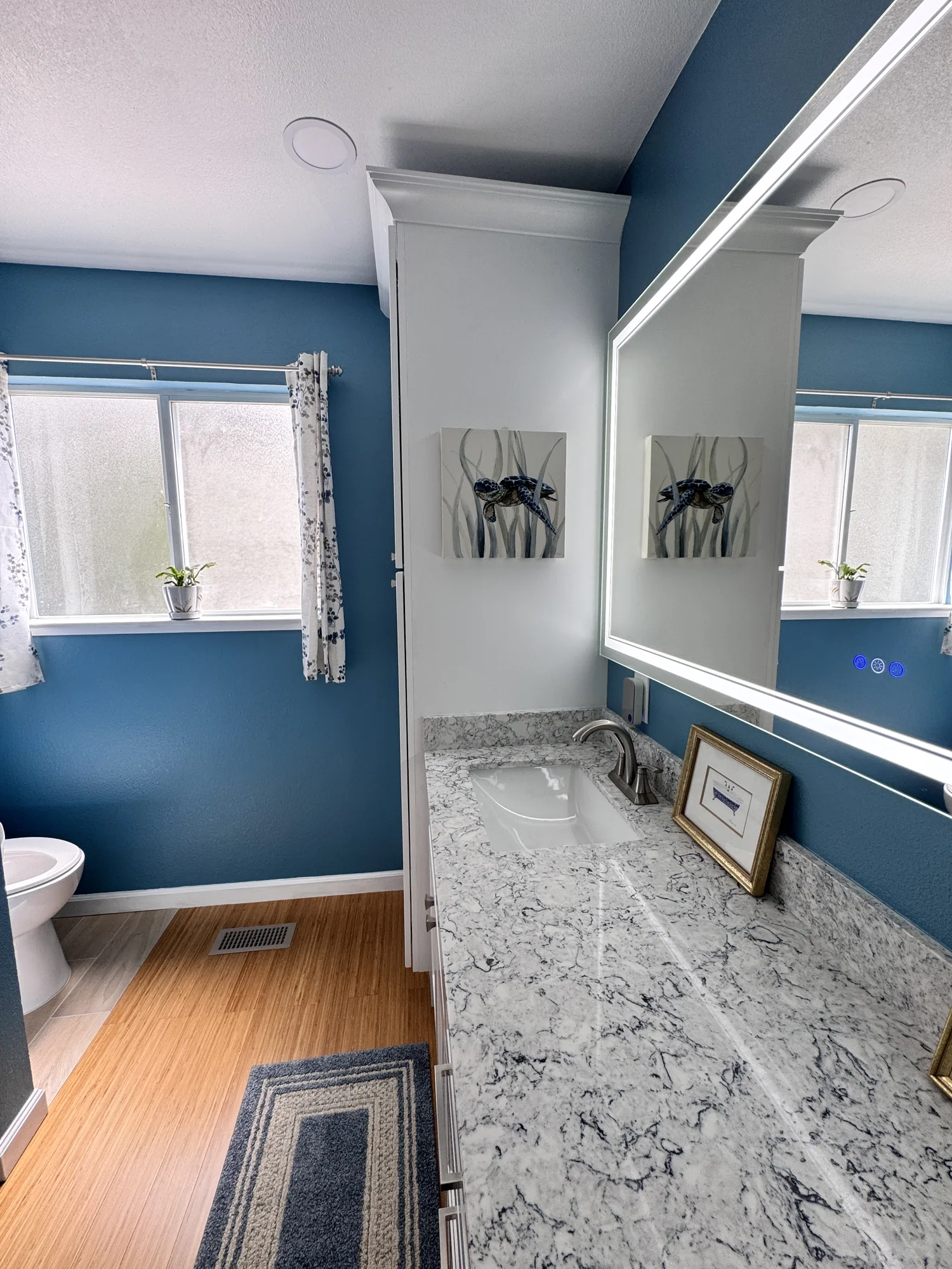 Full bathroom view with blue walls, curtained window, white linen tower and stone-look vanity top.
