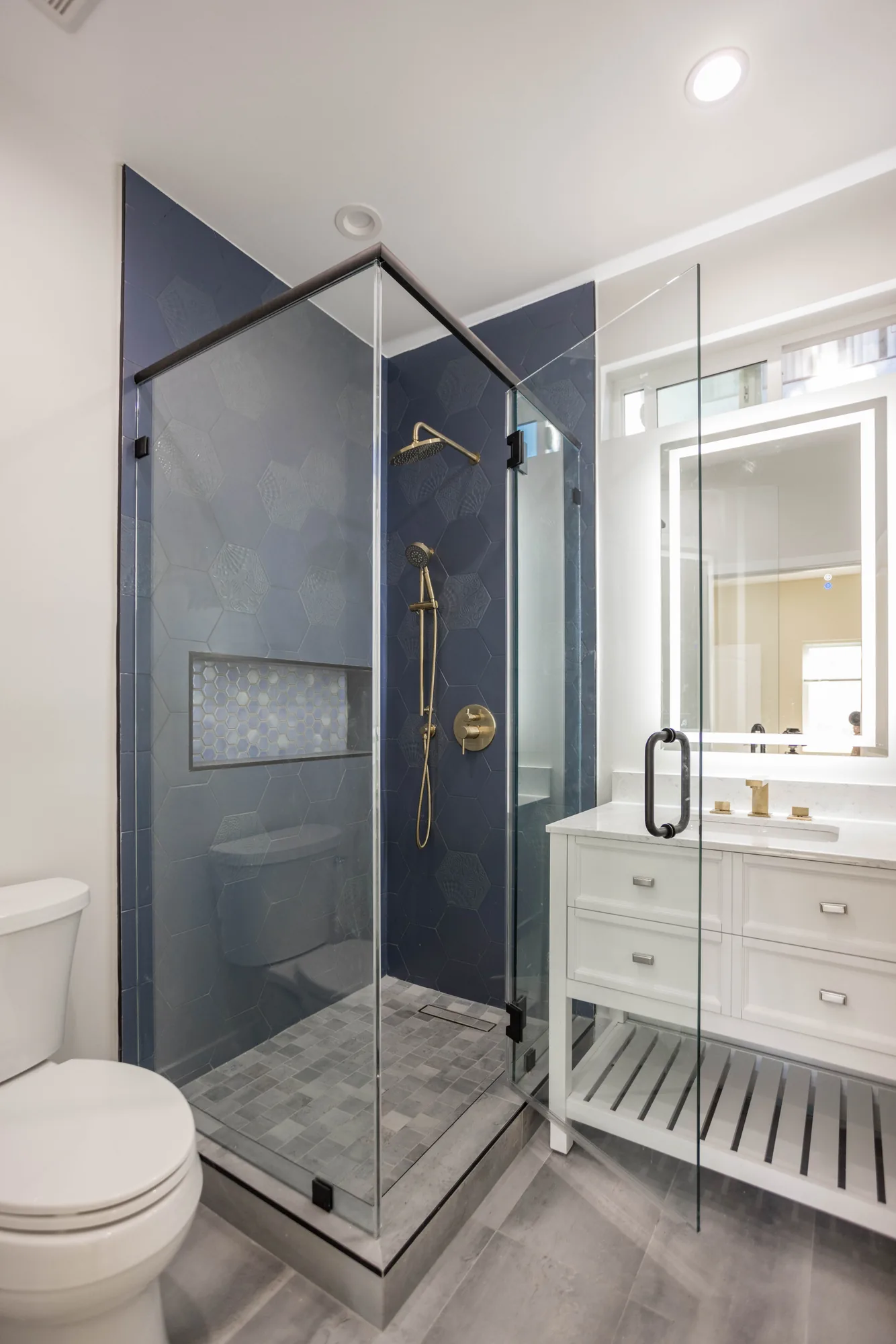 Corner shower in navy hexagon tile with clear glass panels, brass fittings and a gray tile floor.