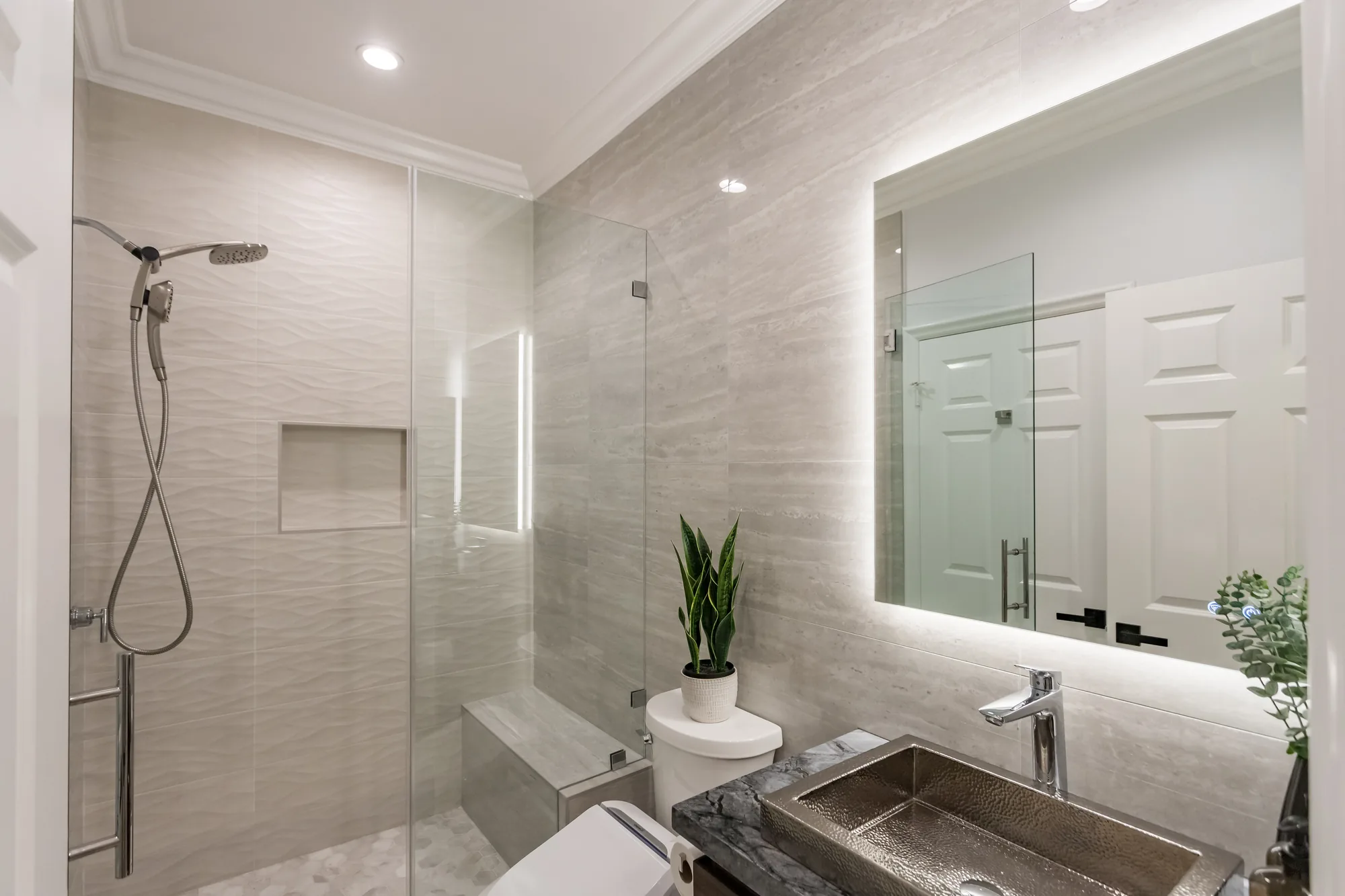 Remodeled bathroom with glass walk-in shower, backlit mirror and hammered metal sink on a stone vanity top.