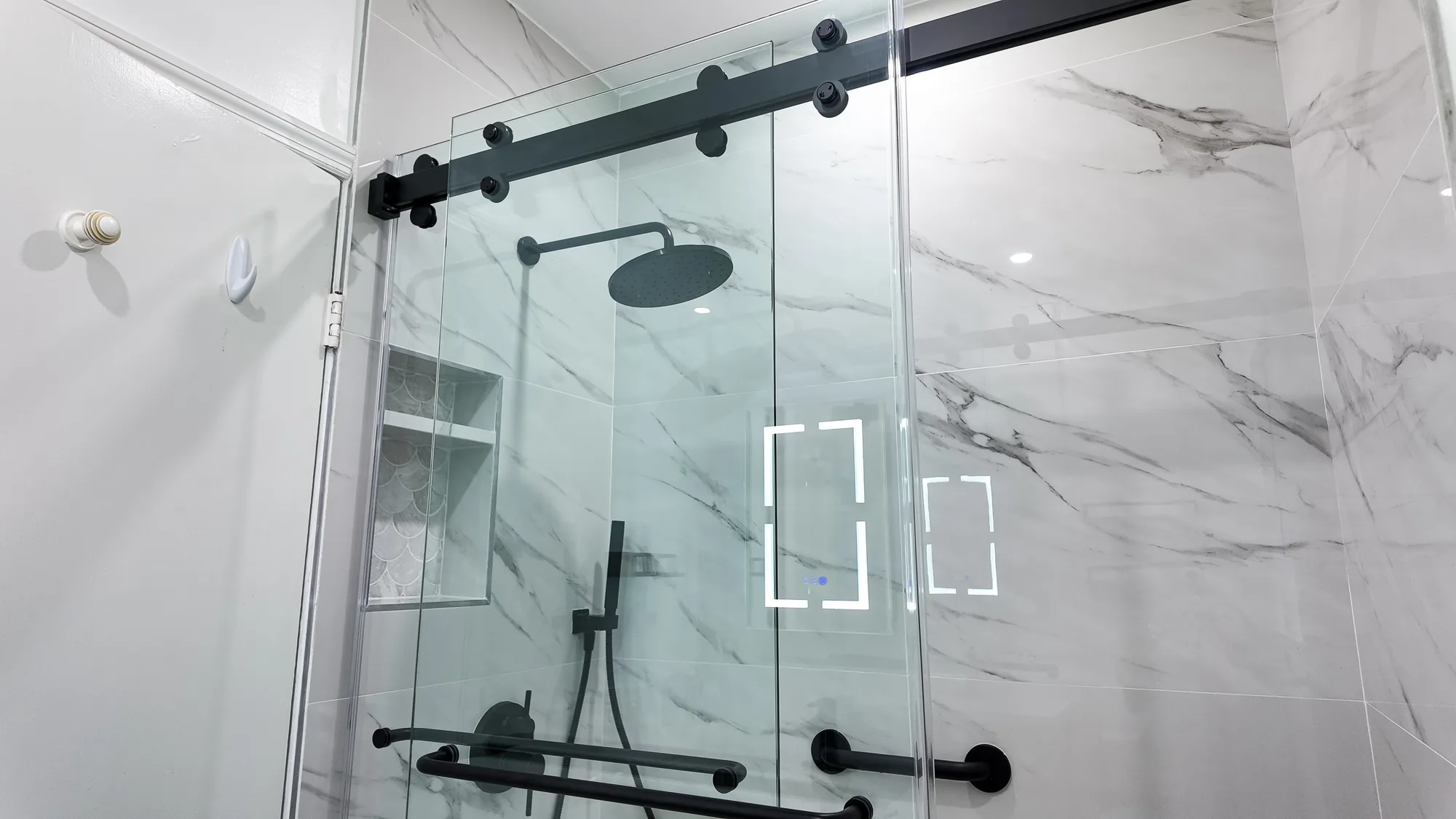 Marble-look tiled shower with sliding glass doors, matte black grab bars, rain head and recessed niche.