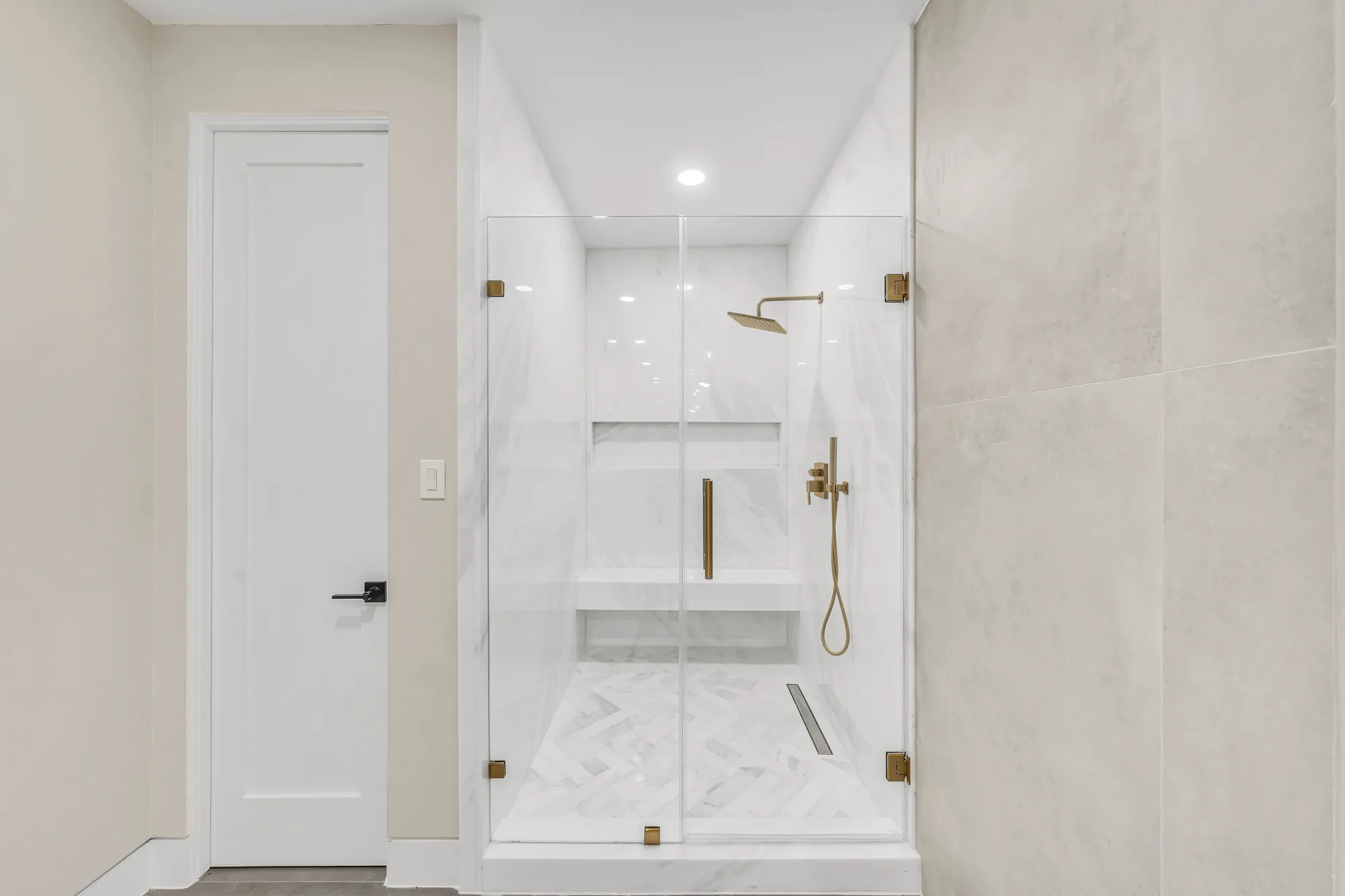 Glass-enclosed shower with marble-look tile, built-in bench, herringbone floor and brushed gold fixtures.