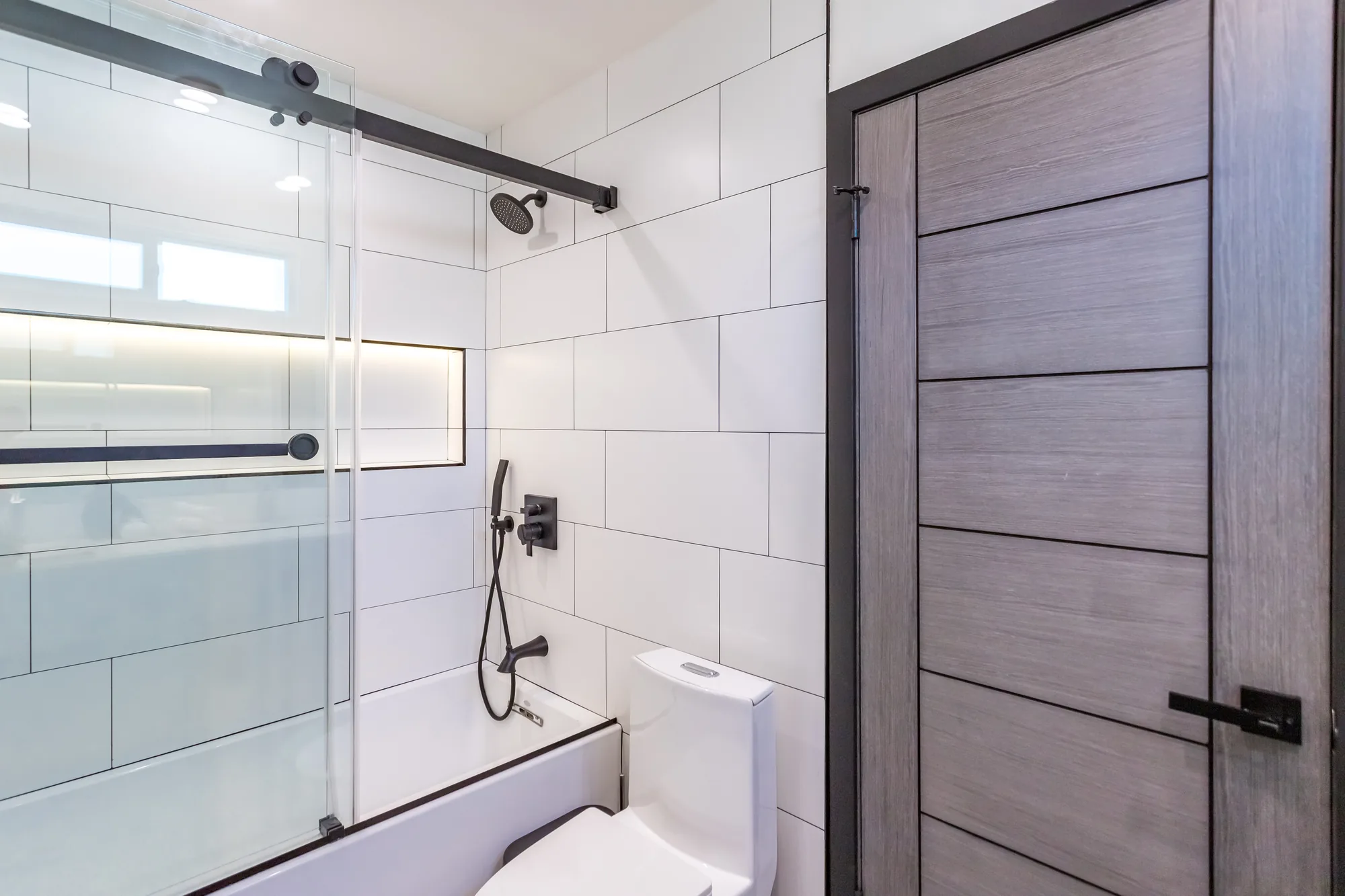 Sliding glass tub enclosure on a black track beside a gray woodgrain door with a black frame.