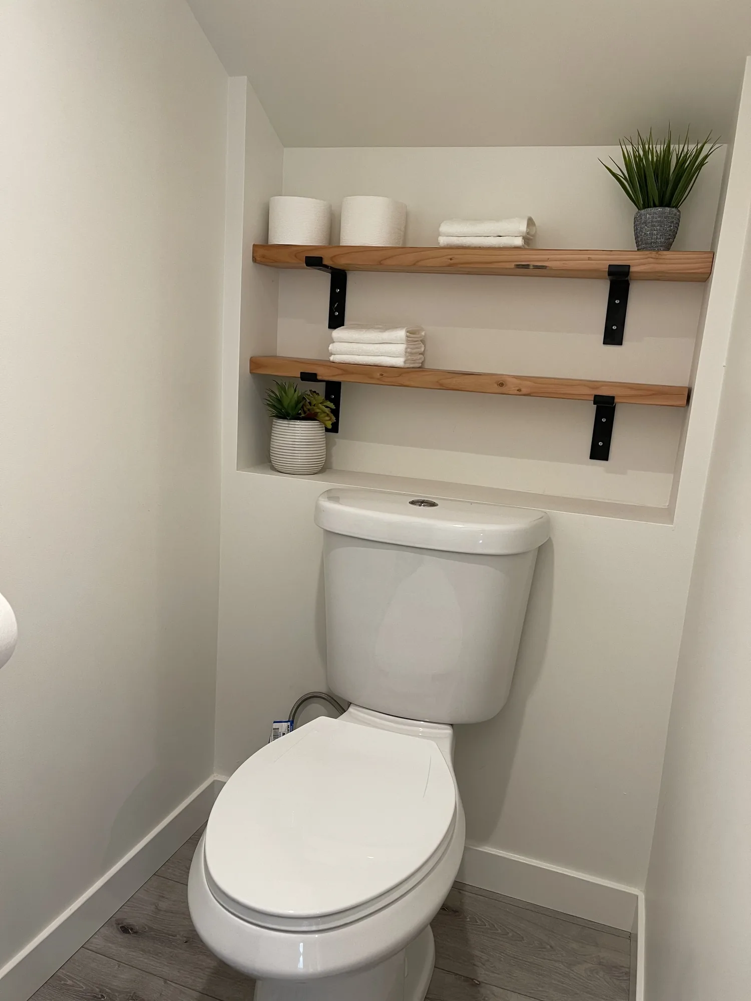 Toilet alcove with two recessed floating wood shelves on black brackets holding towels and plants.