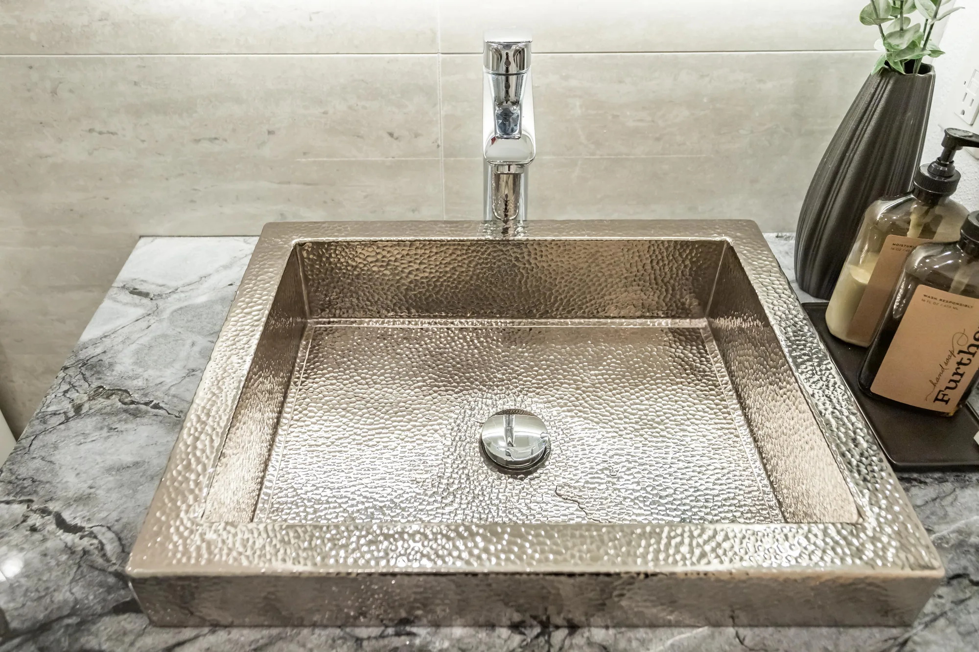 Close-up of a hammered metal vessel sink with a tall chrome faucet on a grey veined stone countertop.