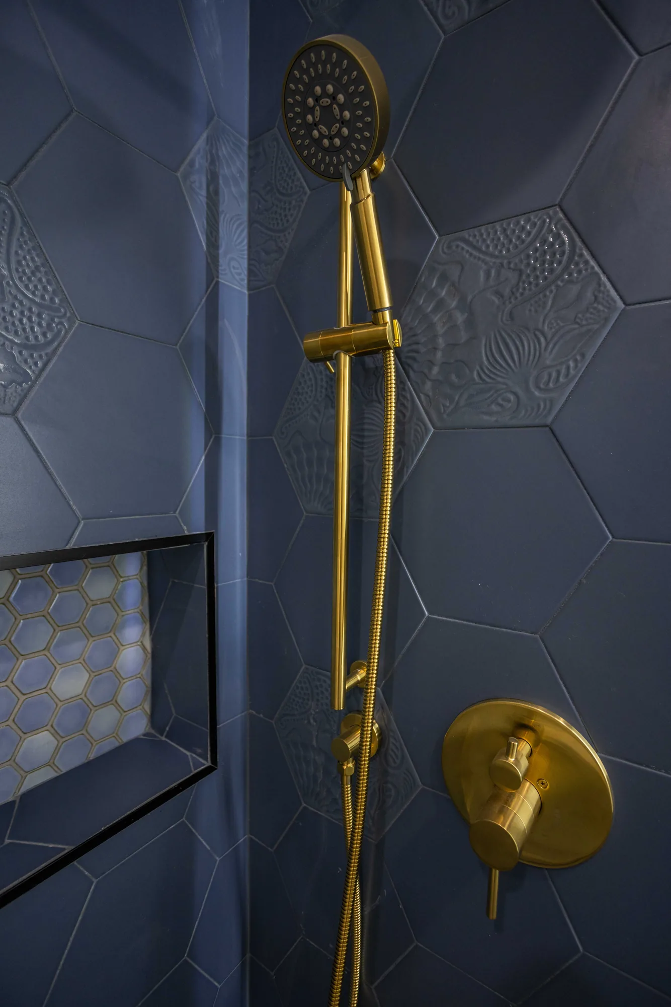 Close view of a brass handheld shower on a slide bar and round valve trim on navy hex tile.