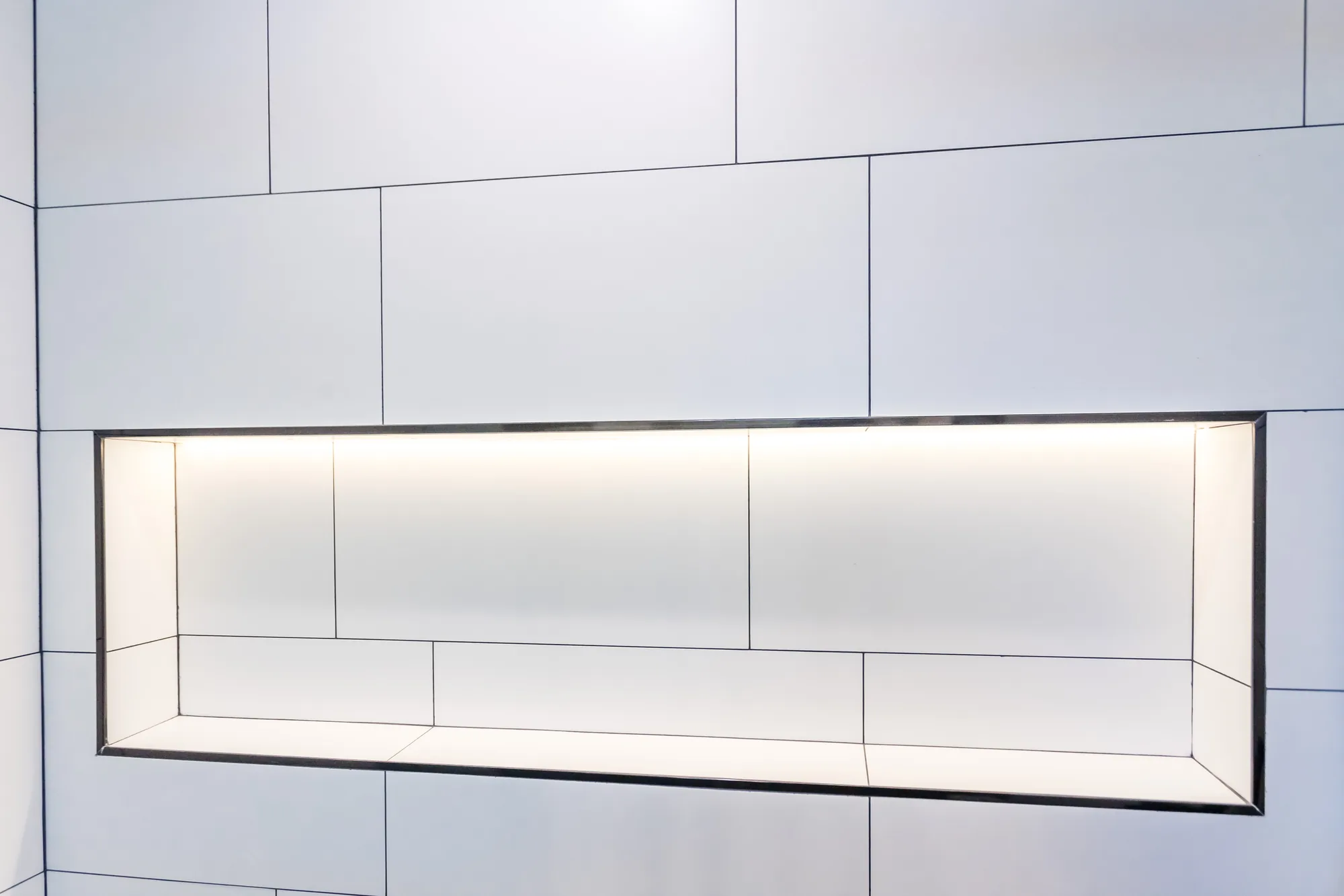 Close-up of a long recessed shower niche with LED lighting set into white large-format tile.