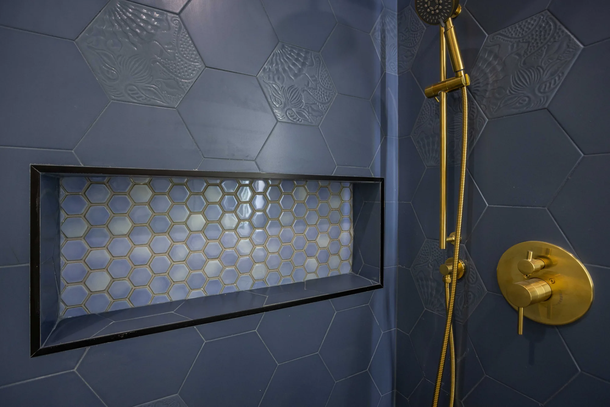 Shower niche lined with iridescent hexagon mosaic and framed in black metal against navy tile.