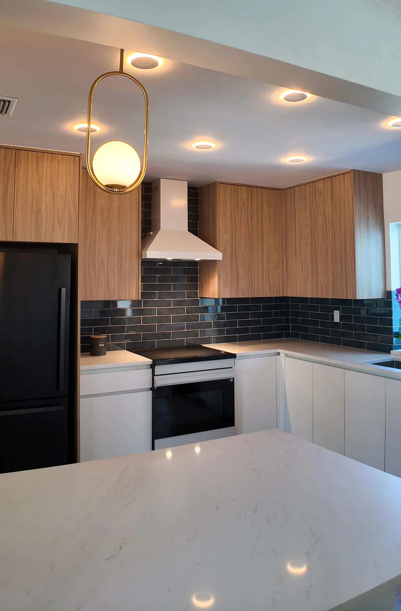 Warmly lit kitchen seen across a light quartz peninsula toward the range wall and dark tile backsplash.