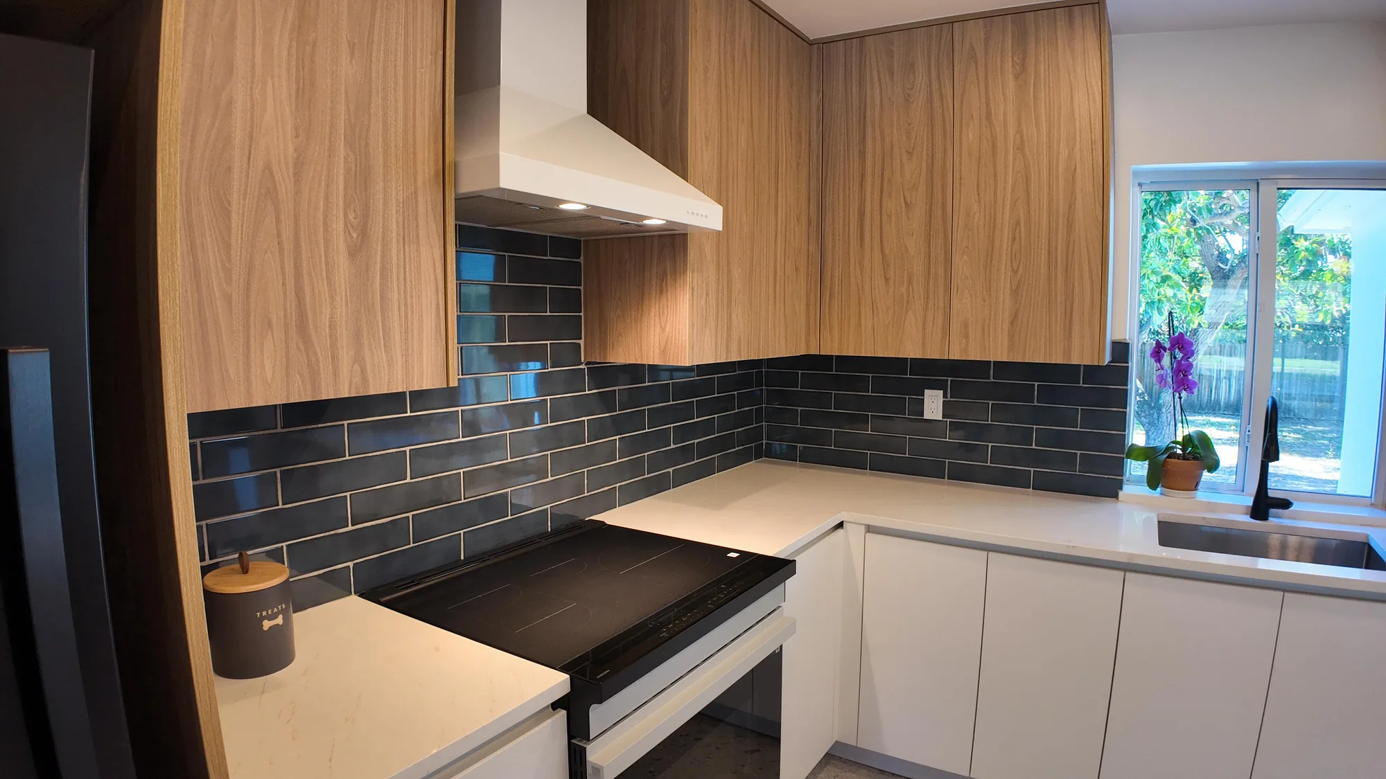Kitchen corner with white chimney hood, dark subway tile backsplash and wood veneer upper cabinets.