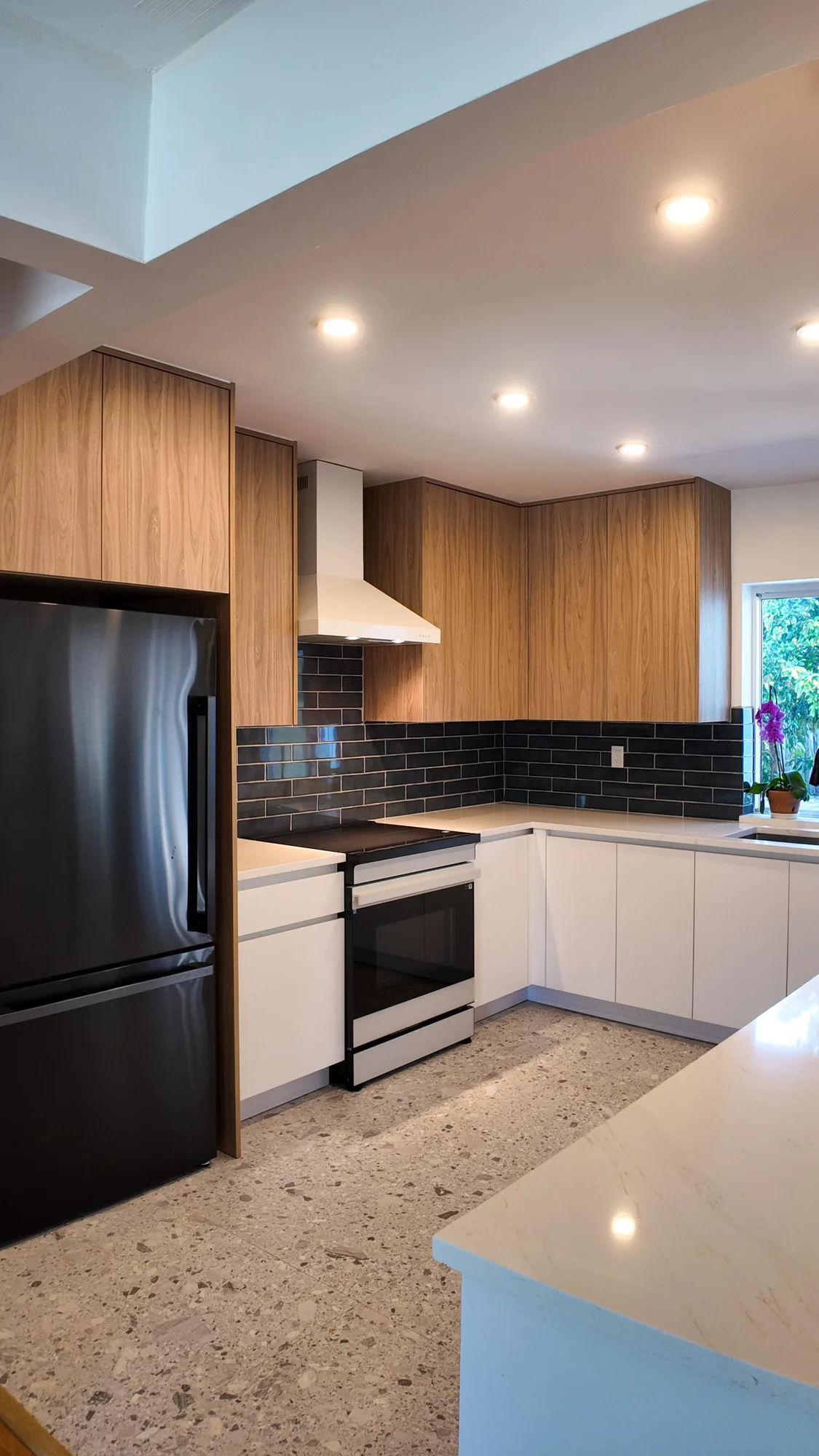 Kitchen with a black refrigerator set into a wood cabinet alcove above a speckled terrazzo-look floor.