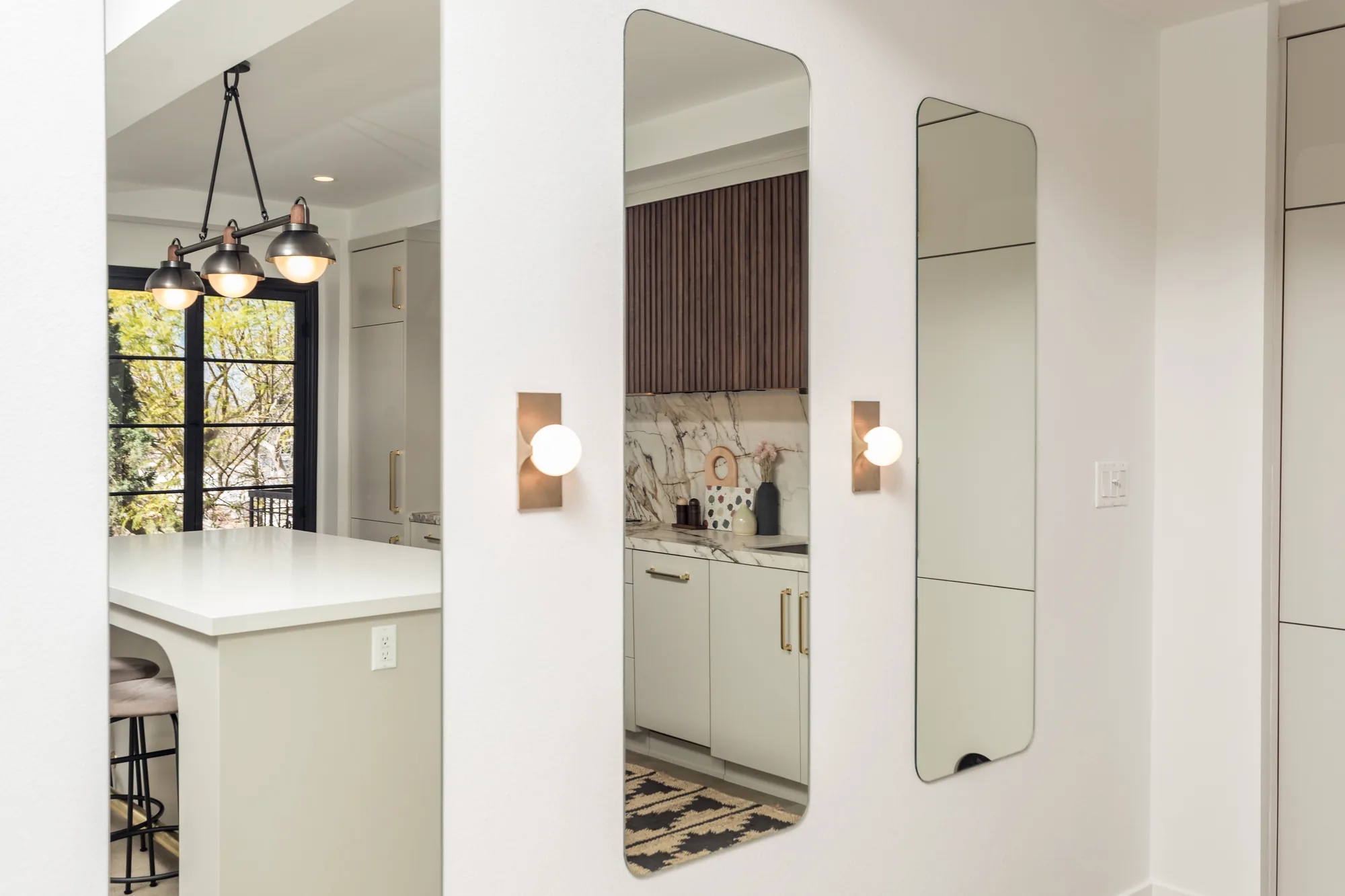 Kitchen and hallway view with pendant lights over a white island, marble backsplash, wall sconces and mirrors.