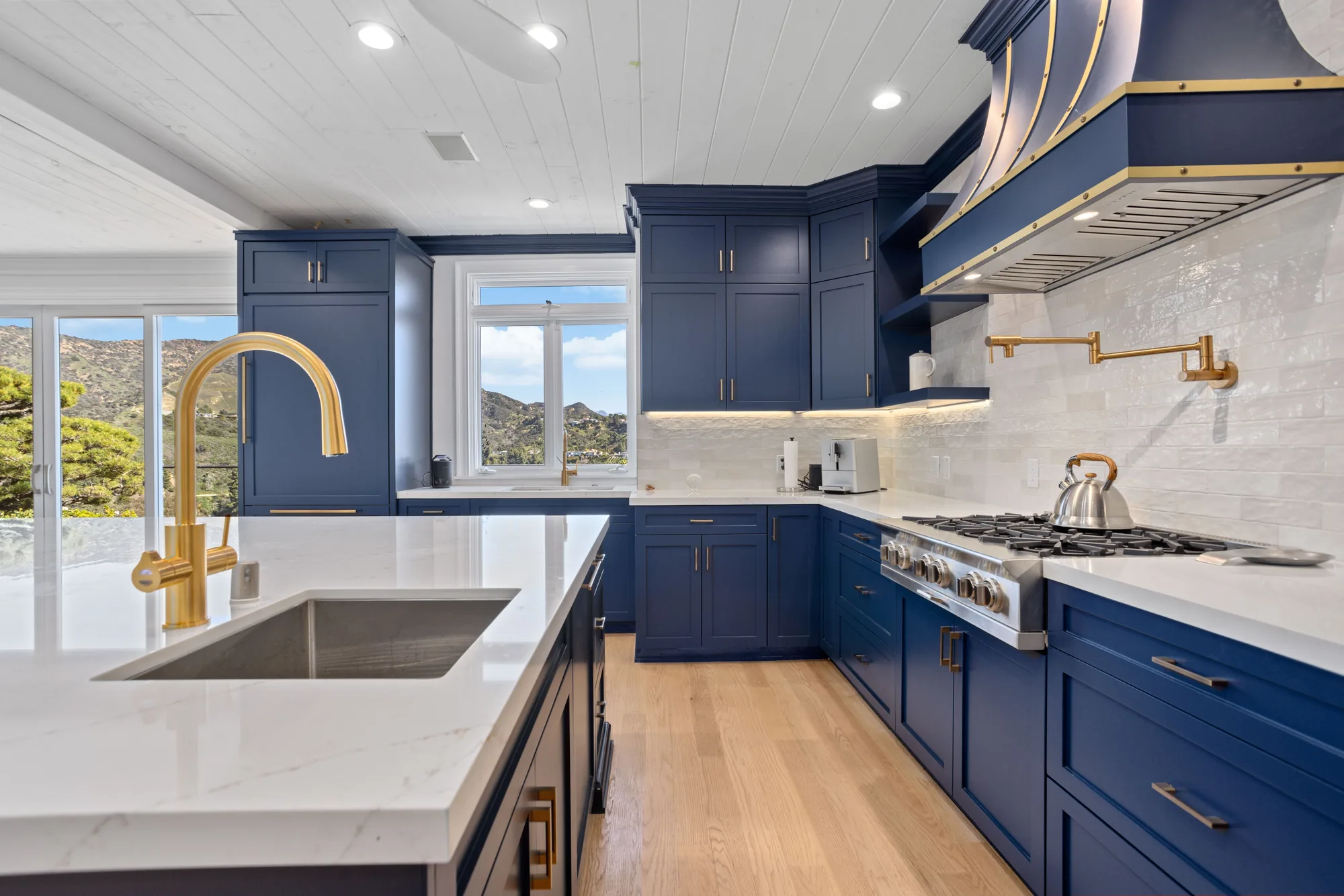 Navy blue kitchen with a brass gooseneck faucet, gas cooktop, marble backsplash and a white quartz island by the window.