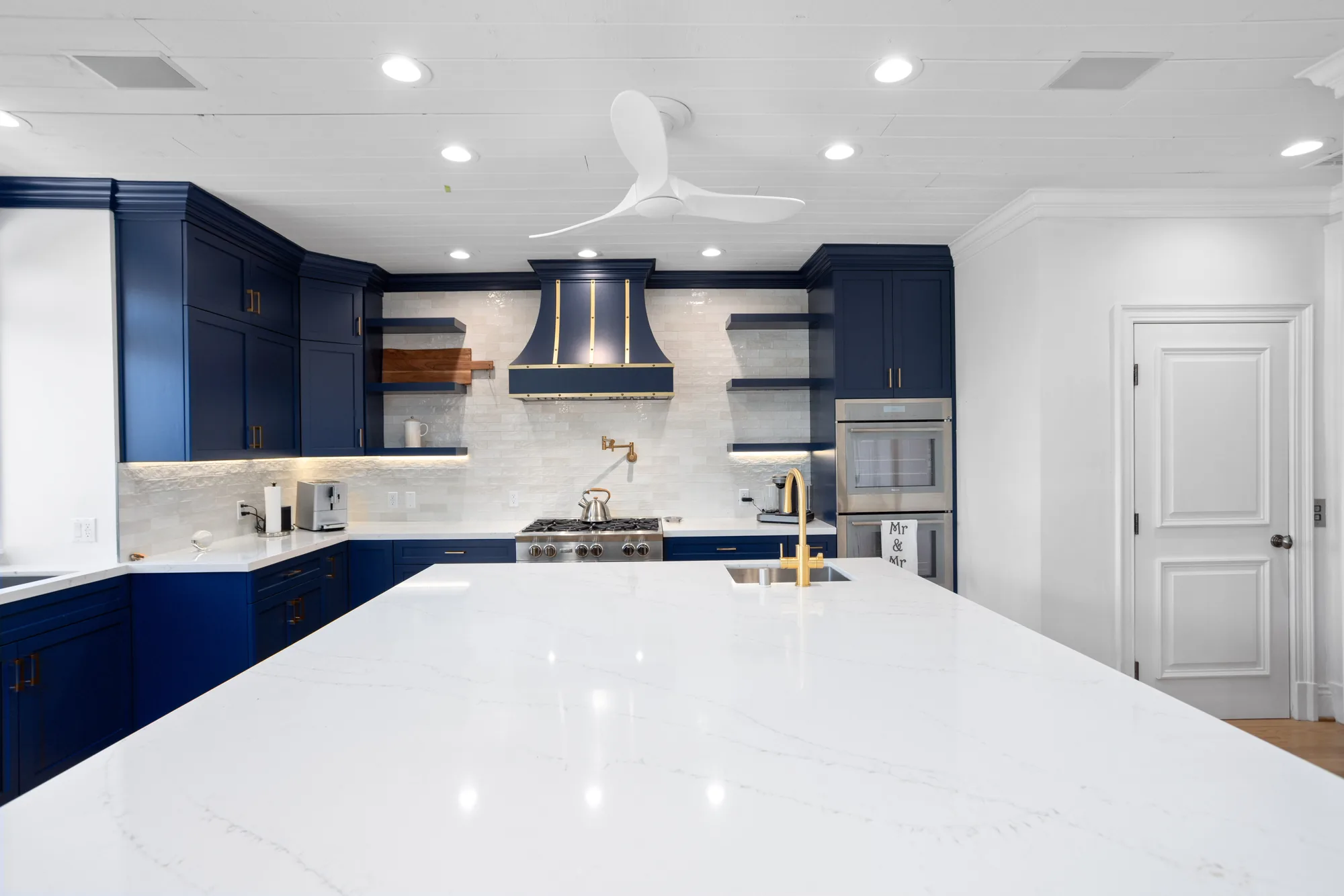 Navy blue kitchen with a large white quartz island, brass-accented range hood, marble backsplash and double wall ovens.