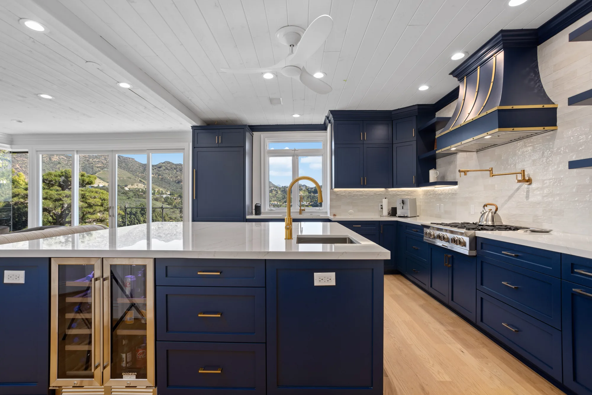 Navy blue kitchen with a white quartz island, brass-trimmed wine fridge, gas range and mountain views through the windows.