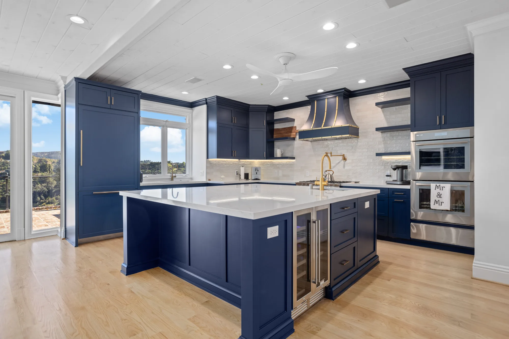 Navy blue kitchen with a white quartz island, wood-and-brass range hood, double wall ovens and light hardwood floors.