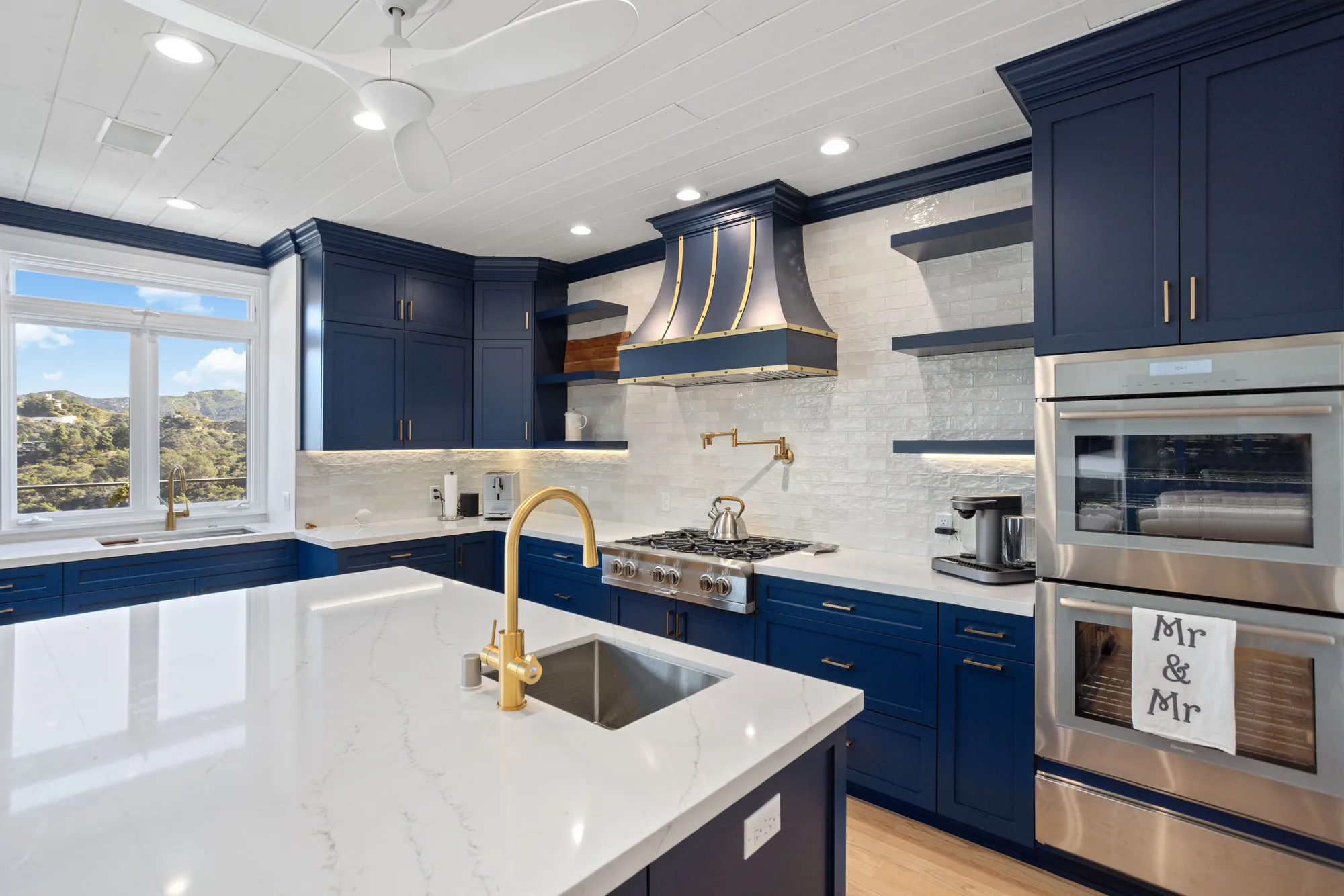 Navy blue kitchen with brass fixtures, gas range, marble backsplash and a white quartz island beneath a hillside window.