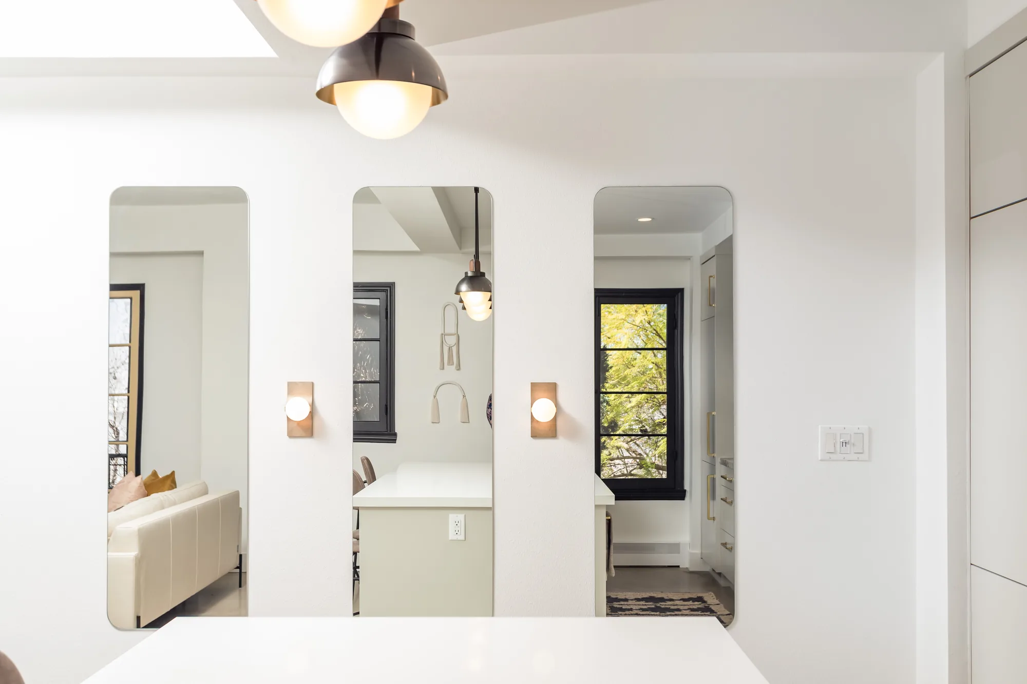 Reflected views of the kitchen and living room in three full-length mirrors with wall sconces and a pendant light.