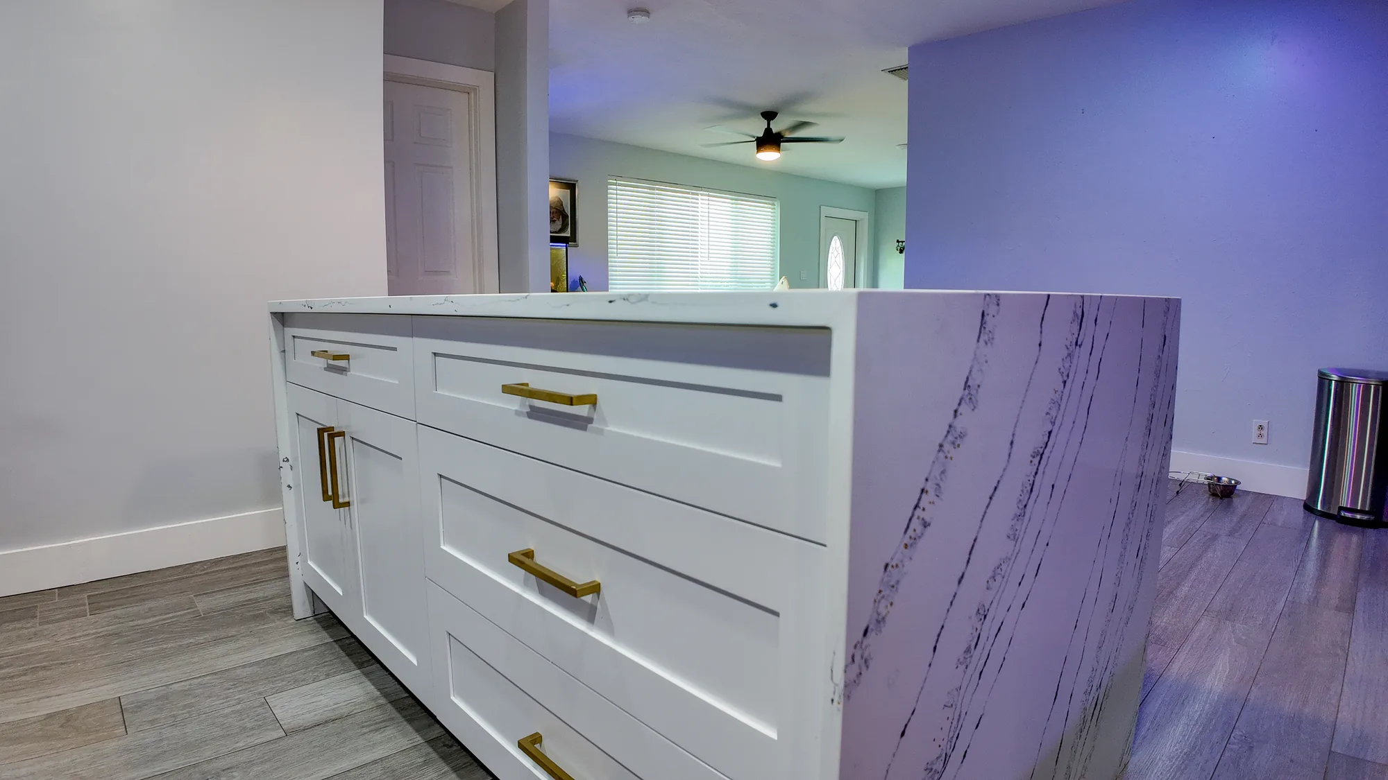 White kitchen island with a marble waterfall edge and gold hardware, open to a living area.