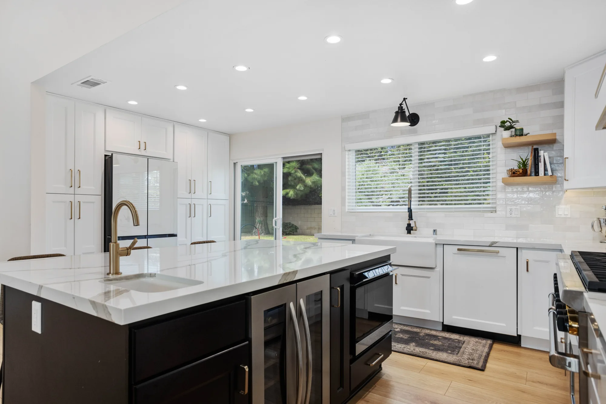 White kitchen with a dark island housing a wine fridge and microwave, brass faucet, apron sink and marble backsplash.