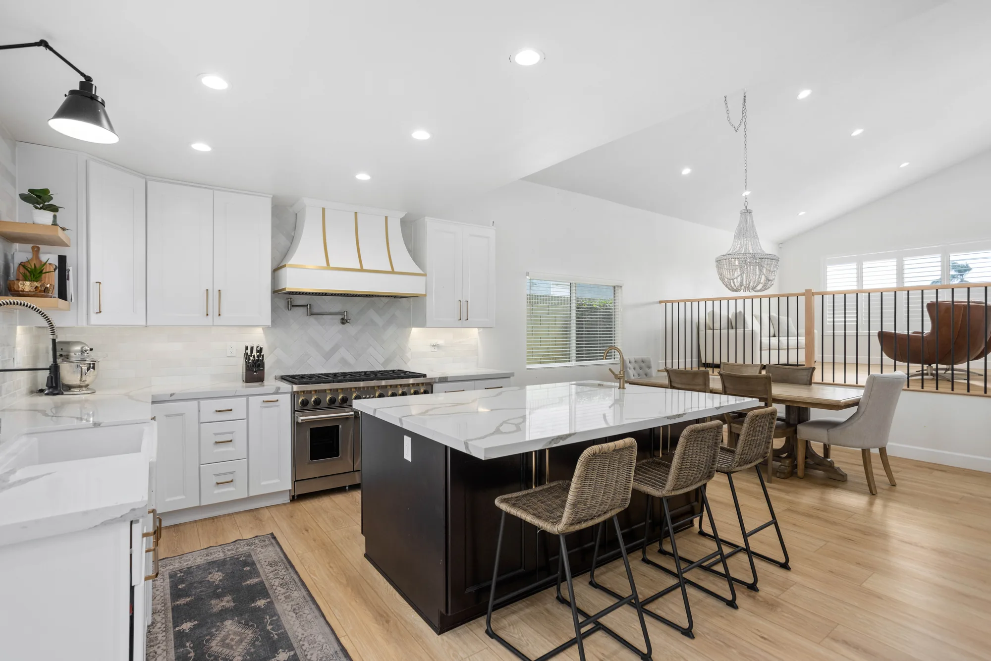 White kitchen with a dark island, marble waterfall counter, brass-strapped hood, herringbone backsplash and rattan stools.