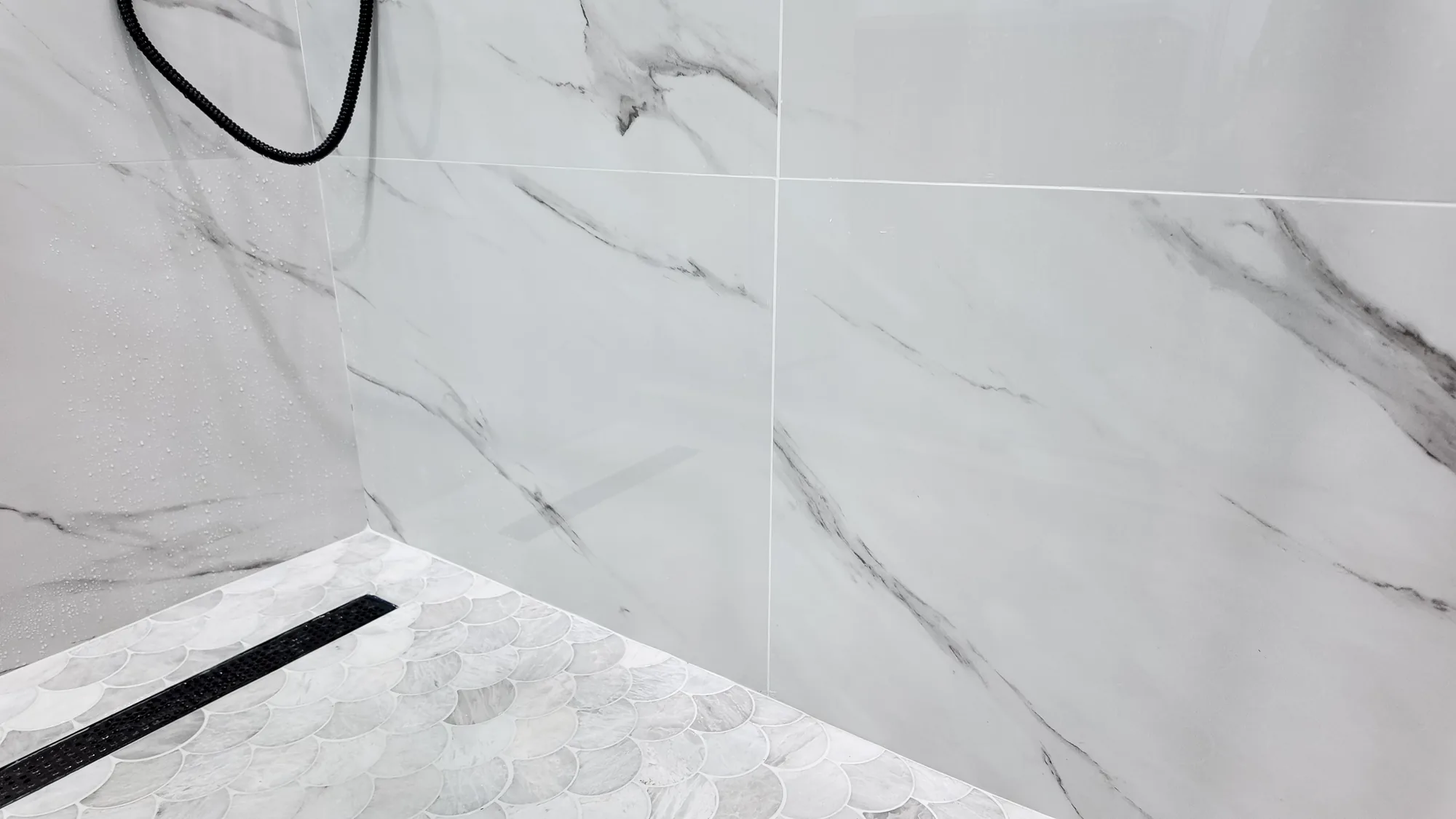 Large-format marble-look wall tile meeting a scalloped mosaic shower pan with a black linear drain.