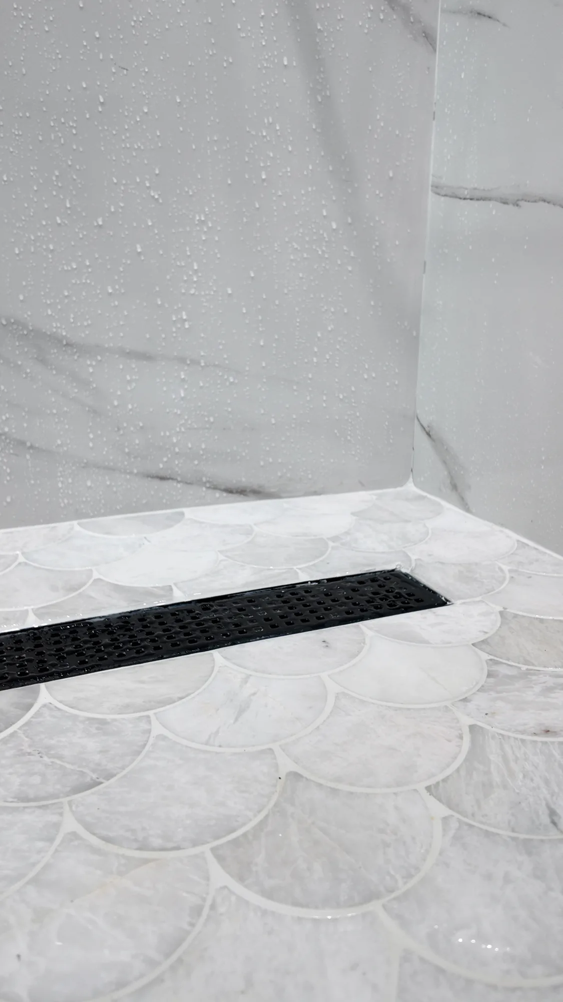 Black linear shower drain set into a scalloped stone mosaic floor beside marble-look wall tile.